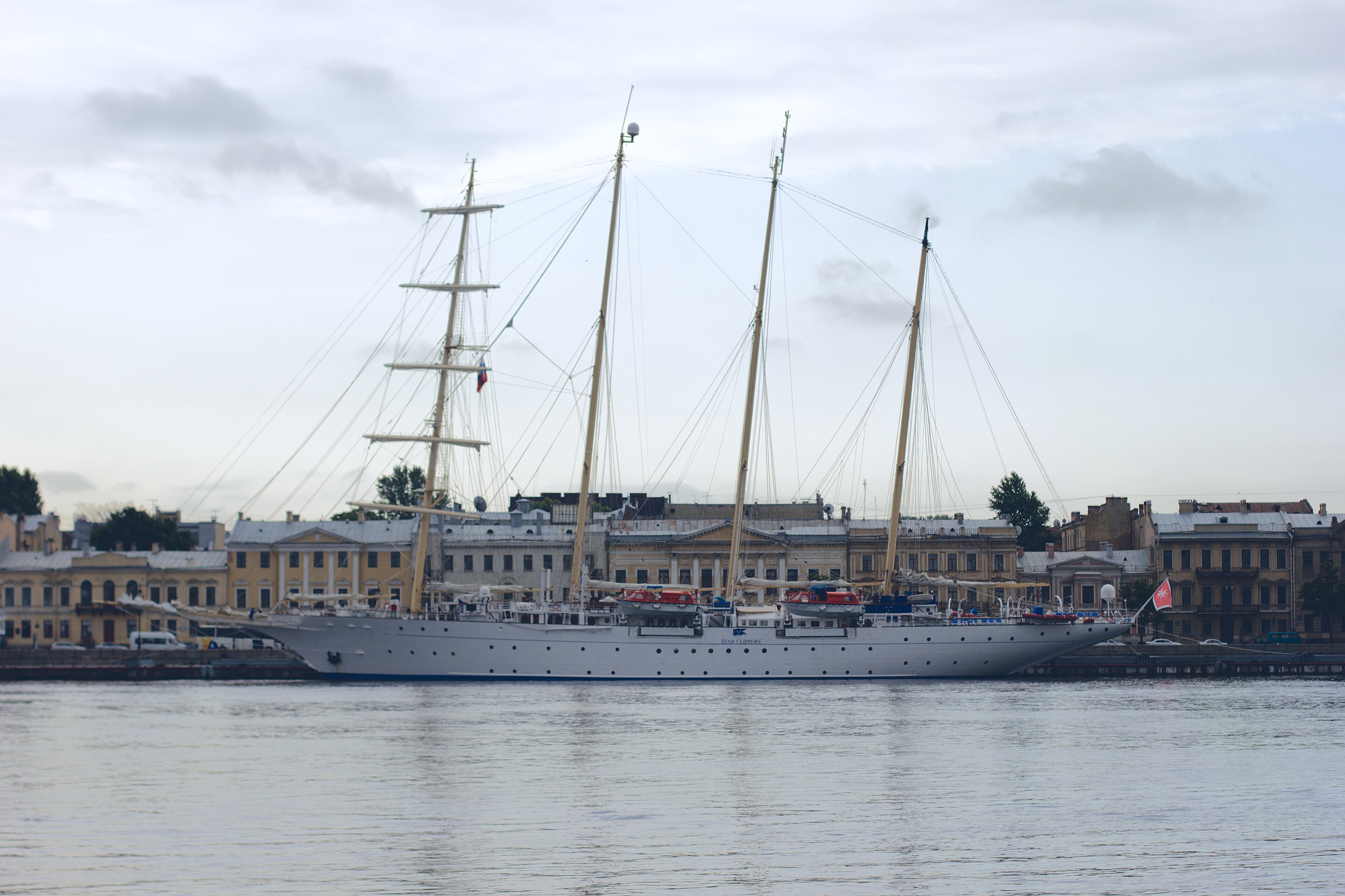Ein großes weißes Segelschiff mit mehreren Masten liegt im Wasser vor einer Stadtkulisse mit historischen Gebäuden.