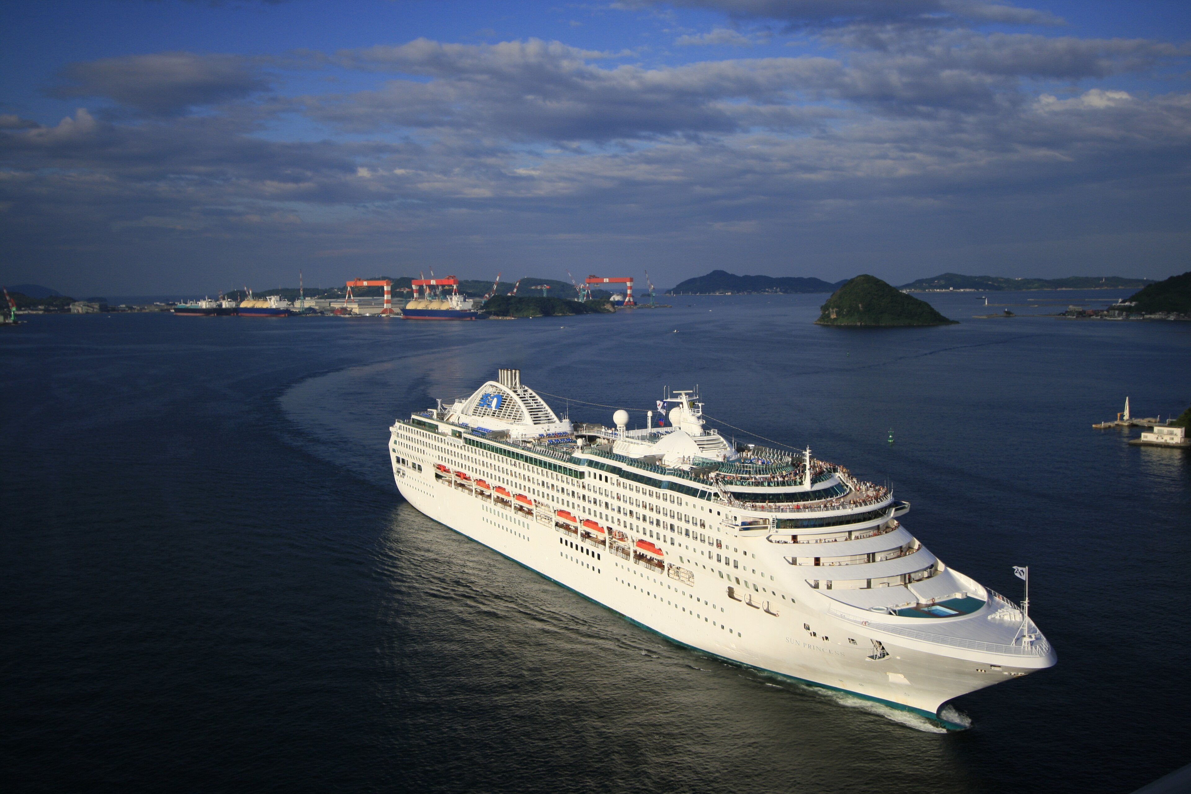 Ein großes weißes Kreuzfahrtschiff fährt auf dem Meer in der Nähe einer Schiffswerft mit Kränen und anderen Schiffen.