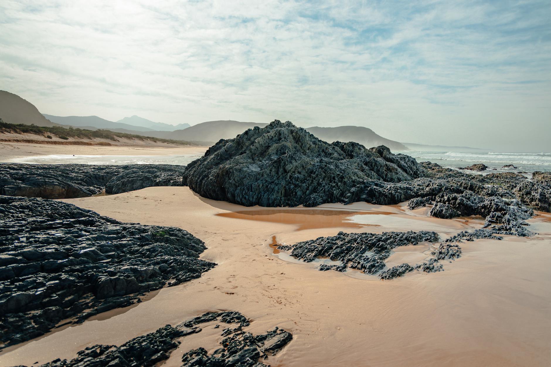 Ein felsiger Strand mit Sand und Felsen im Vordergrund und dem Meer und Bergen im Hintergrund.