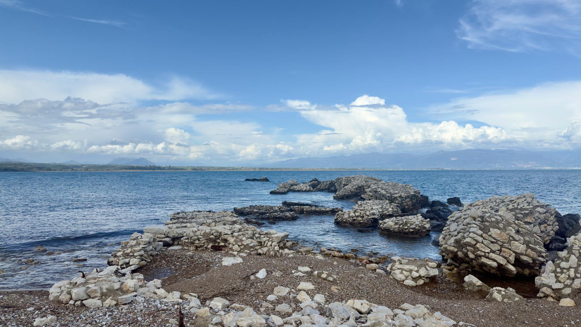Eine felsige Küste mit antiken Ruinen, die ins blaue Meer ragen, unter einem Himmel mit dramatischen Wolken und fernen Bergen.