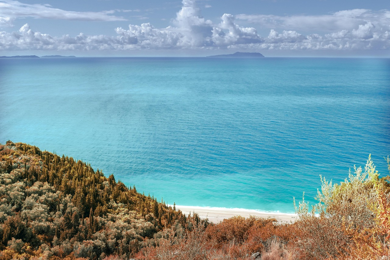 Ein Blick von einem huegeligen Ufer mit Vegetation auf einen weissen Sandstrand und das tuerkisblaue Meer, im Hintergrund eine Insel und Wolken am Himmel.