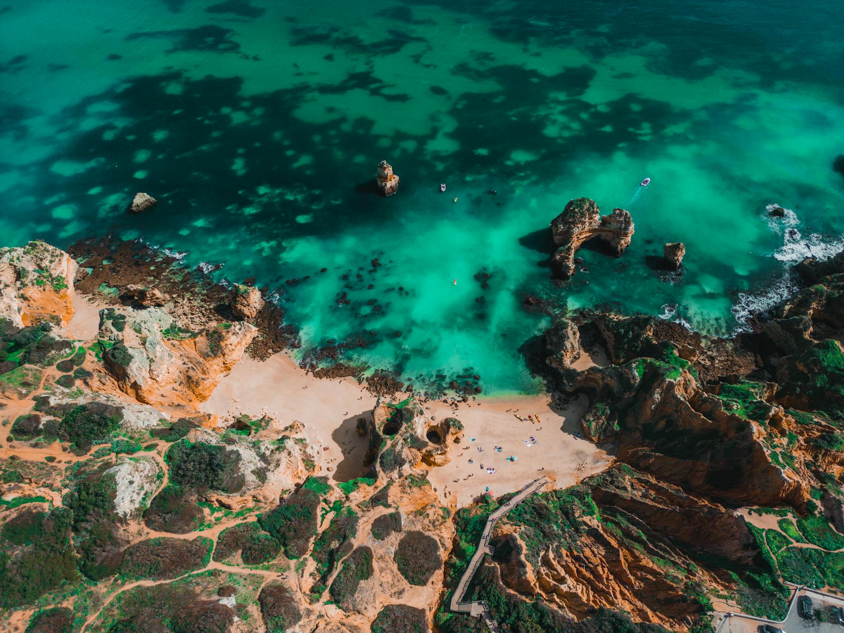 Algarve, Portugal - Luftaufnahme eines Sandstrandes mit felsigen Klippen und tuerkisem Wasser, auf dem mehrere Boote und Menschen zu sehen sind.