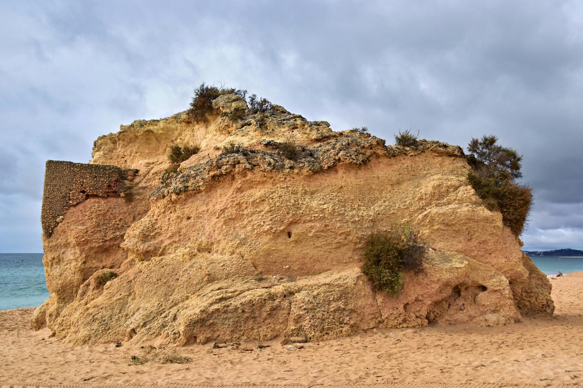 Algarve, Portugal - Eine ockerfarbene Felsformation mit Vegetation und einer steinernen Mauer an einem Sandstrand mit dem Meer im Hintergrund.