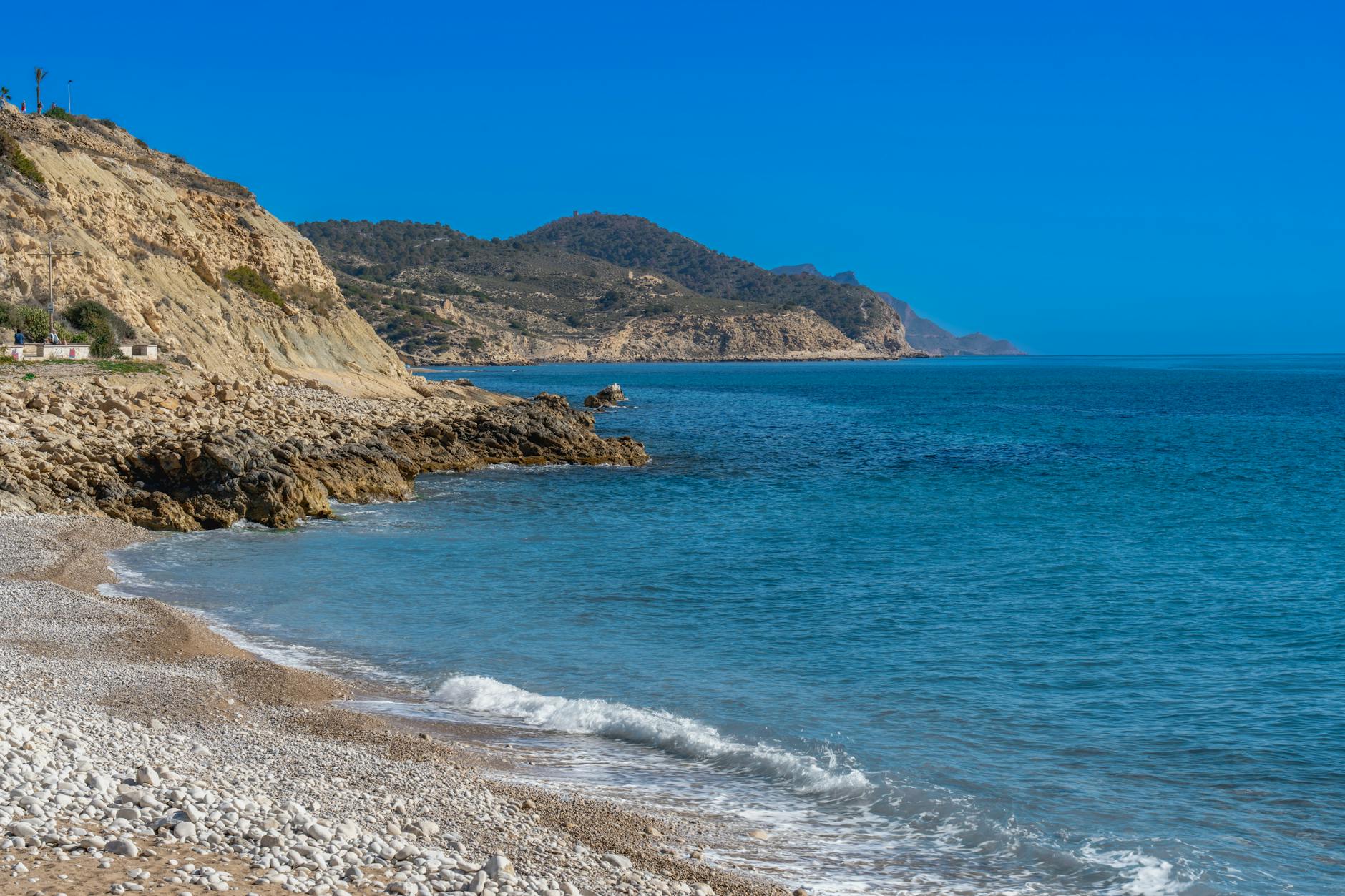 Ein Kiesstrand mit felsiger Kueste und tuerkisem Meer unter klarem blauen Himmel.