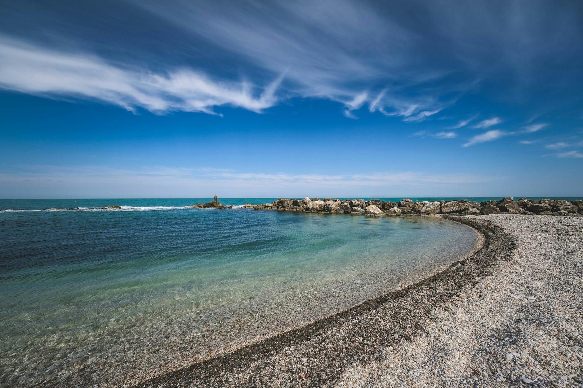 Ein Kiesstrand mit einer Felsmole, die ins Meer ragt, unter einem blauen Himmel mit Schleierwolken.