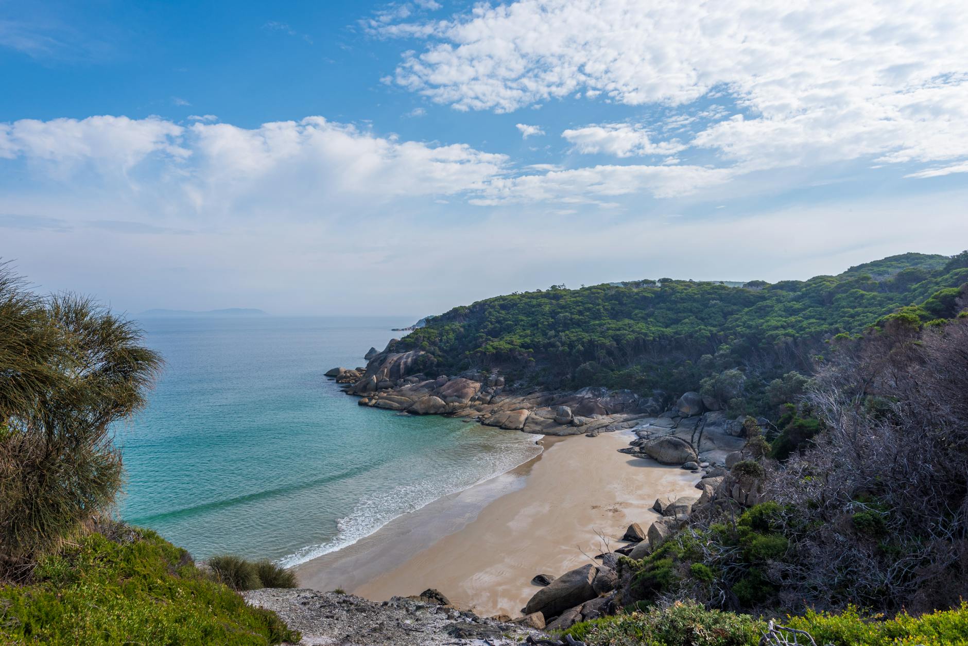 Eine Bucht mit einem Sandstrand, umgeben von felsigen Klippen und gruener Vegetation, mit tuerkisem Wasser und einem wolkenverhangenen Himmel.