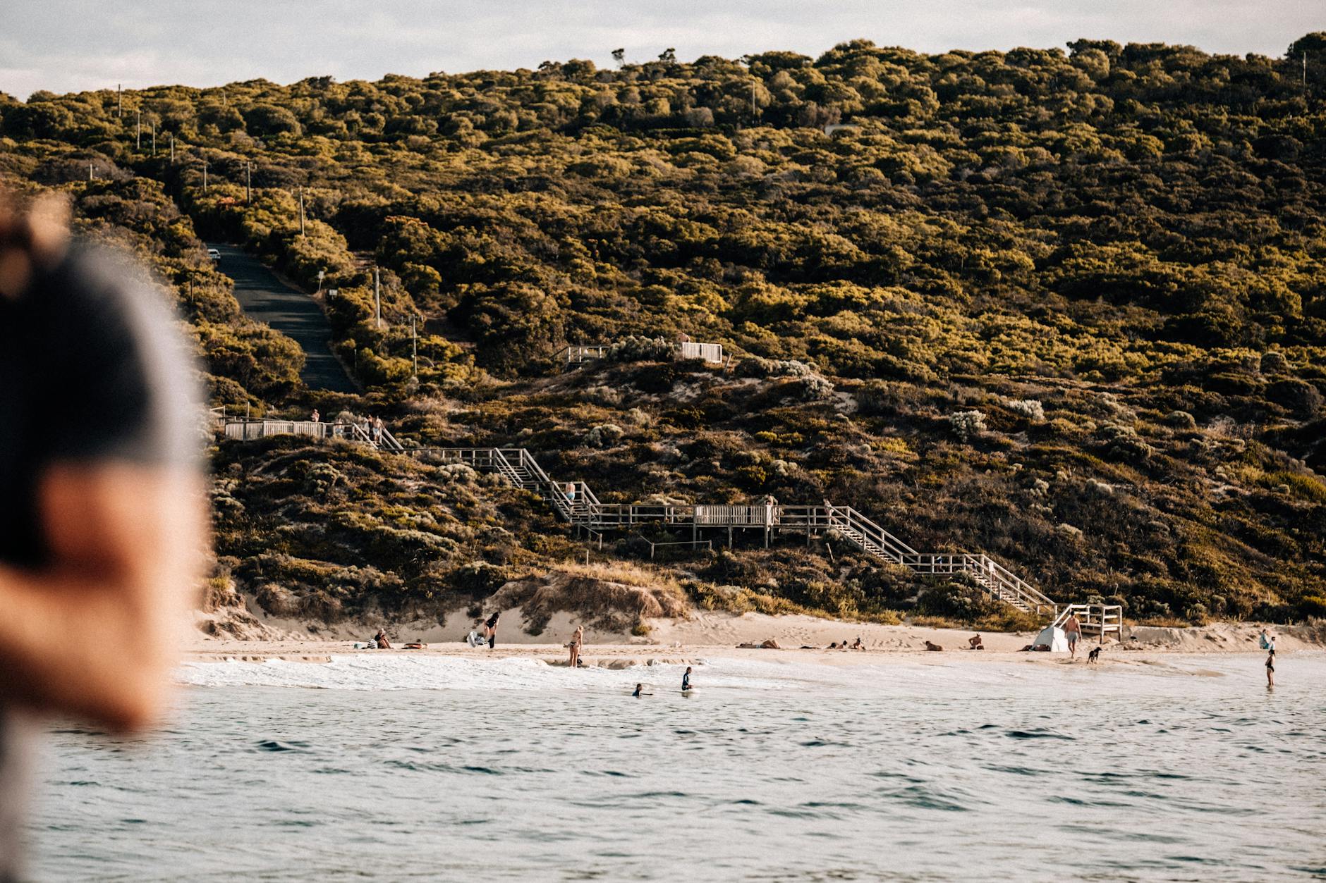 Ein Strand mit duenenbewachsenen Huegeln im Hintergrund, Meer und Menschen am Strand.