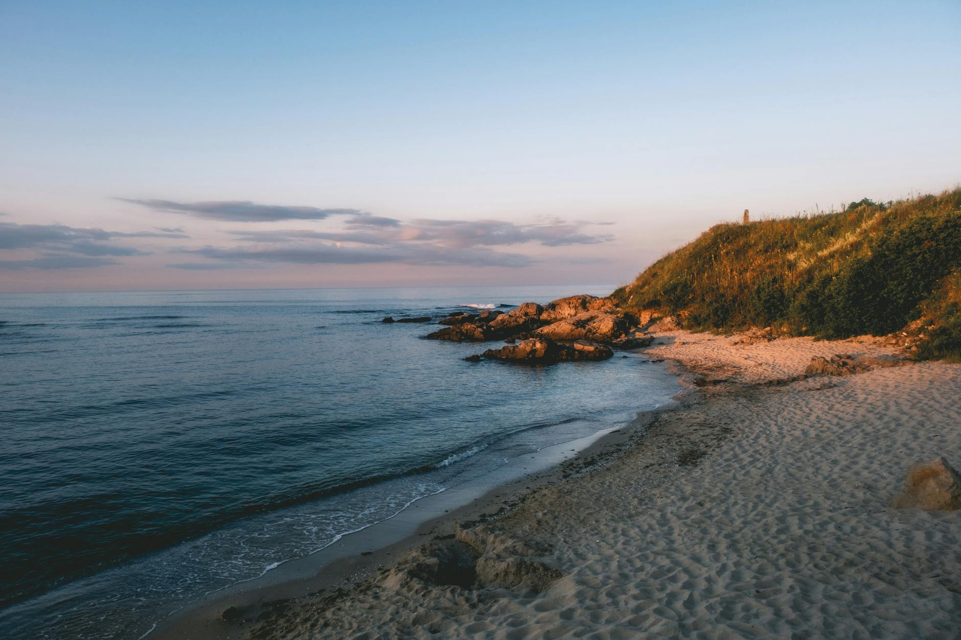 Ein sandiger Strand mit Felsen und einer grasbewachsenen Klippe am Meer bei Sonnenuntergang.