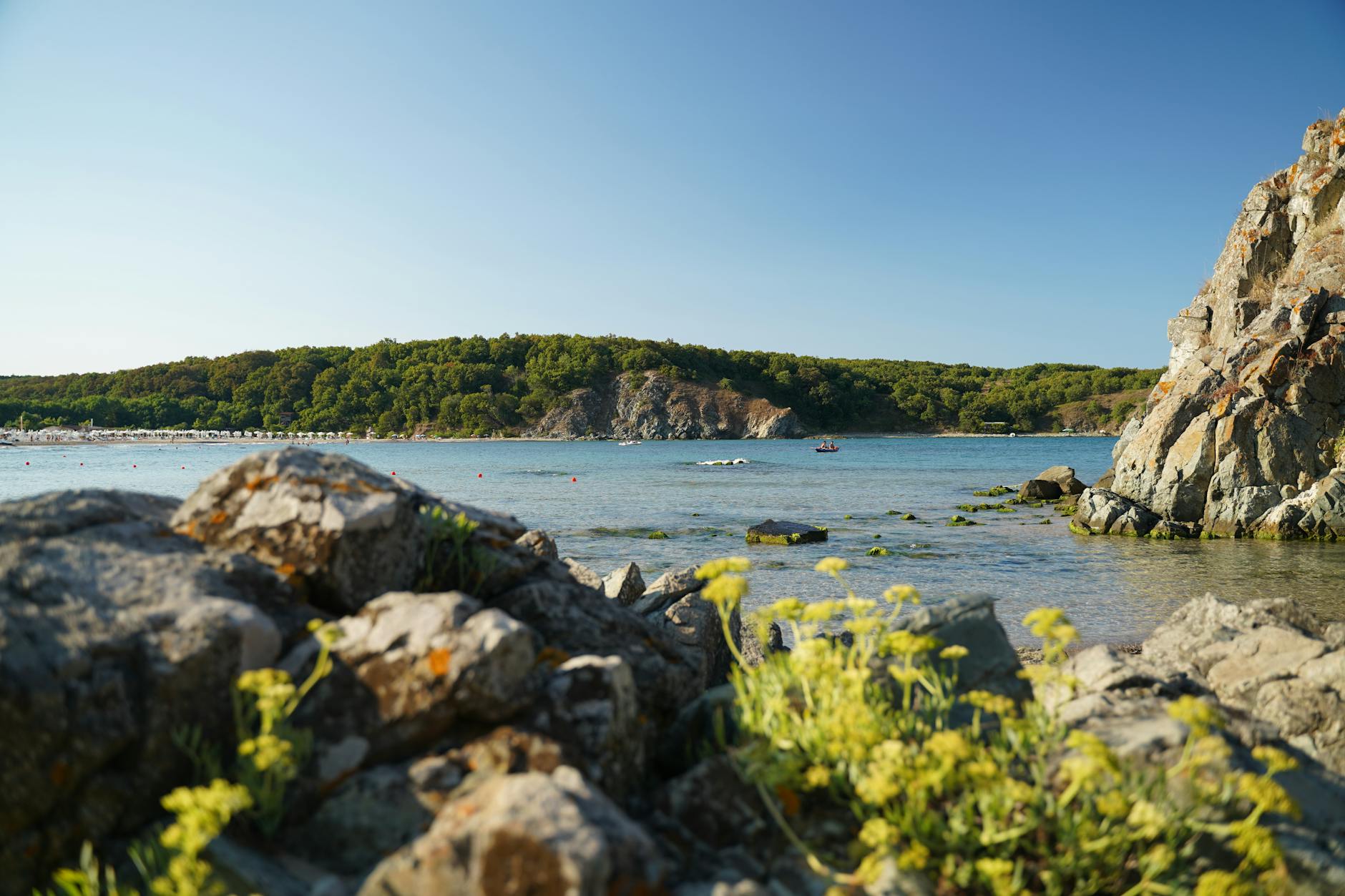 Eine felsige Kueste mit gruenen Huegeln im Hintergrund und einem kleinen Boot auf dem ruhigen Meer.