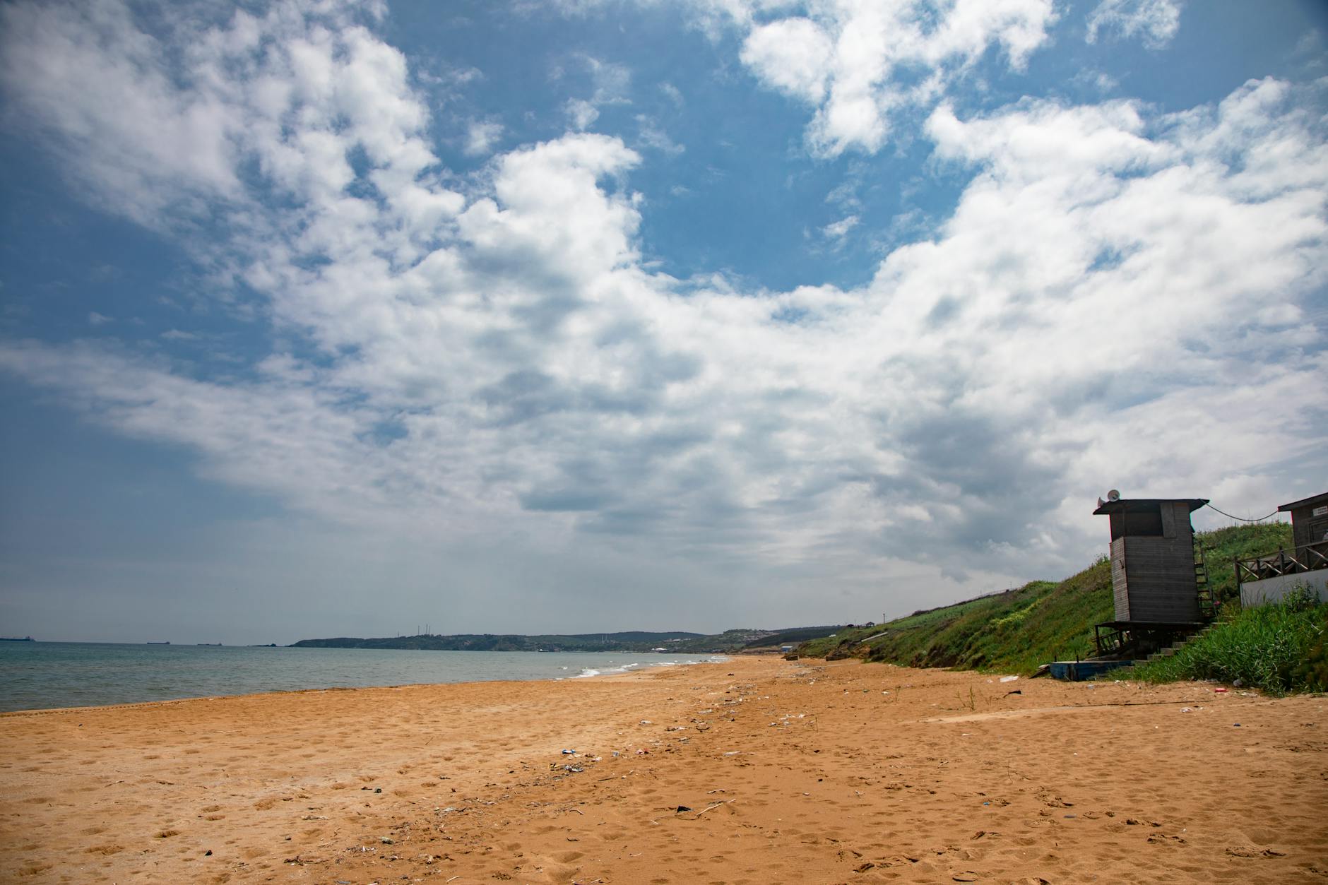 Ein weiter Sandstrand mit einem kleinen Holzhaus auf einer grasbewachsenen Anhoehe im Hintergrund unter einem Himmel mit Wolken.