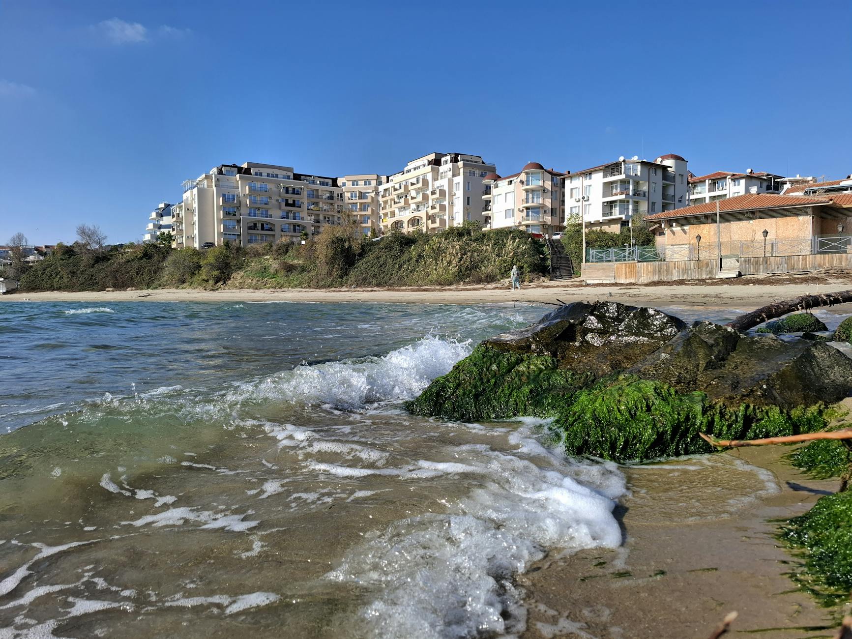 Eine Welle bricht sich an einem felsigen Strand mit Gebäuden im Hintergrund.