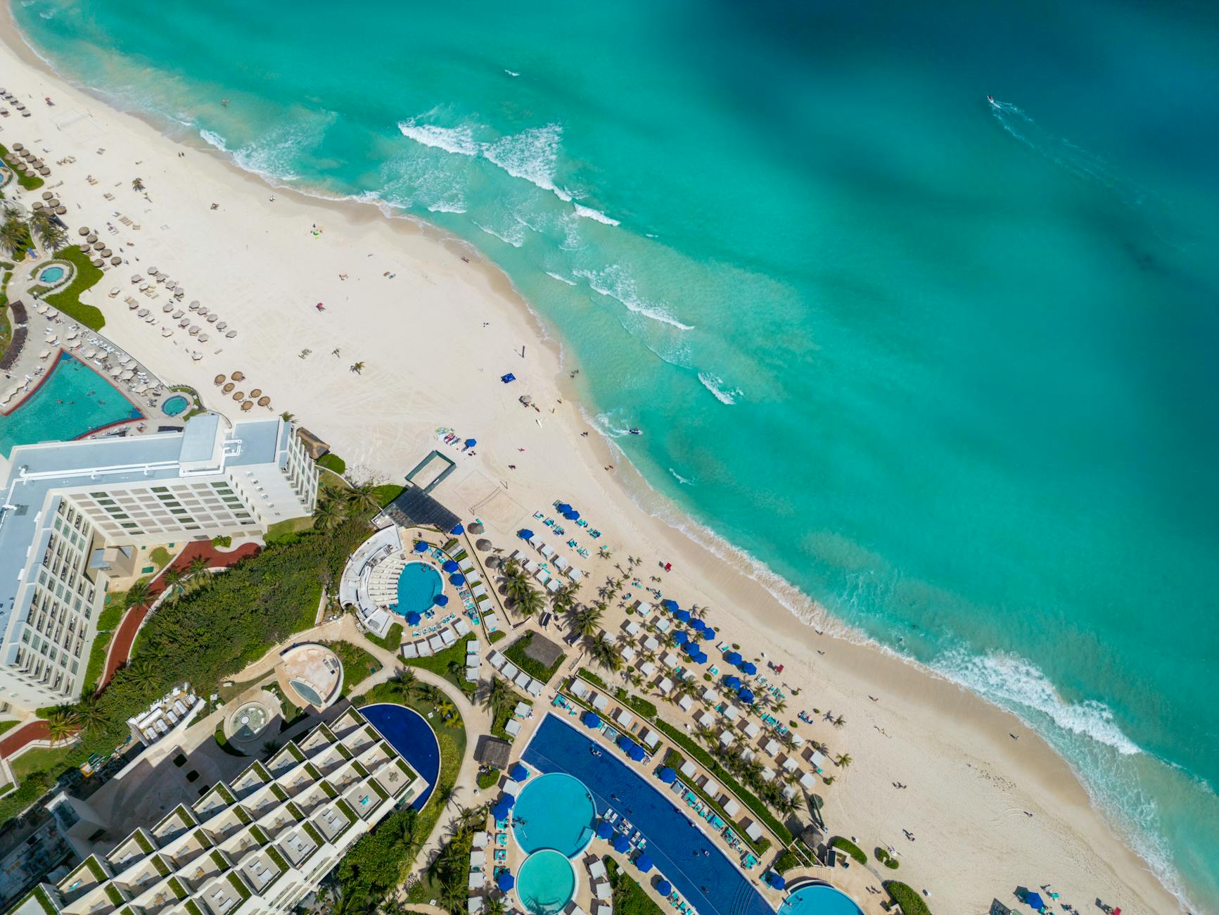 Luftaufnahme einer tropischen Hotelanlage mit mehreren Pools und einem breiten Sandstrand, der in das türkisfarbene Meer übergeht.