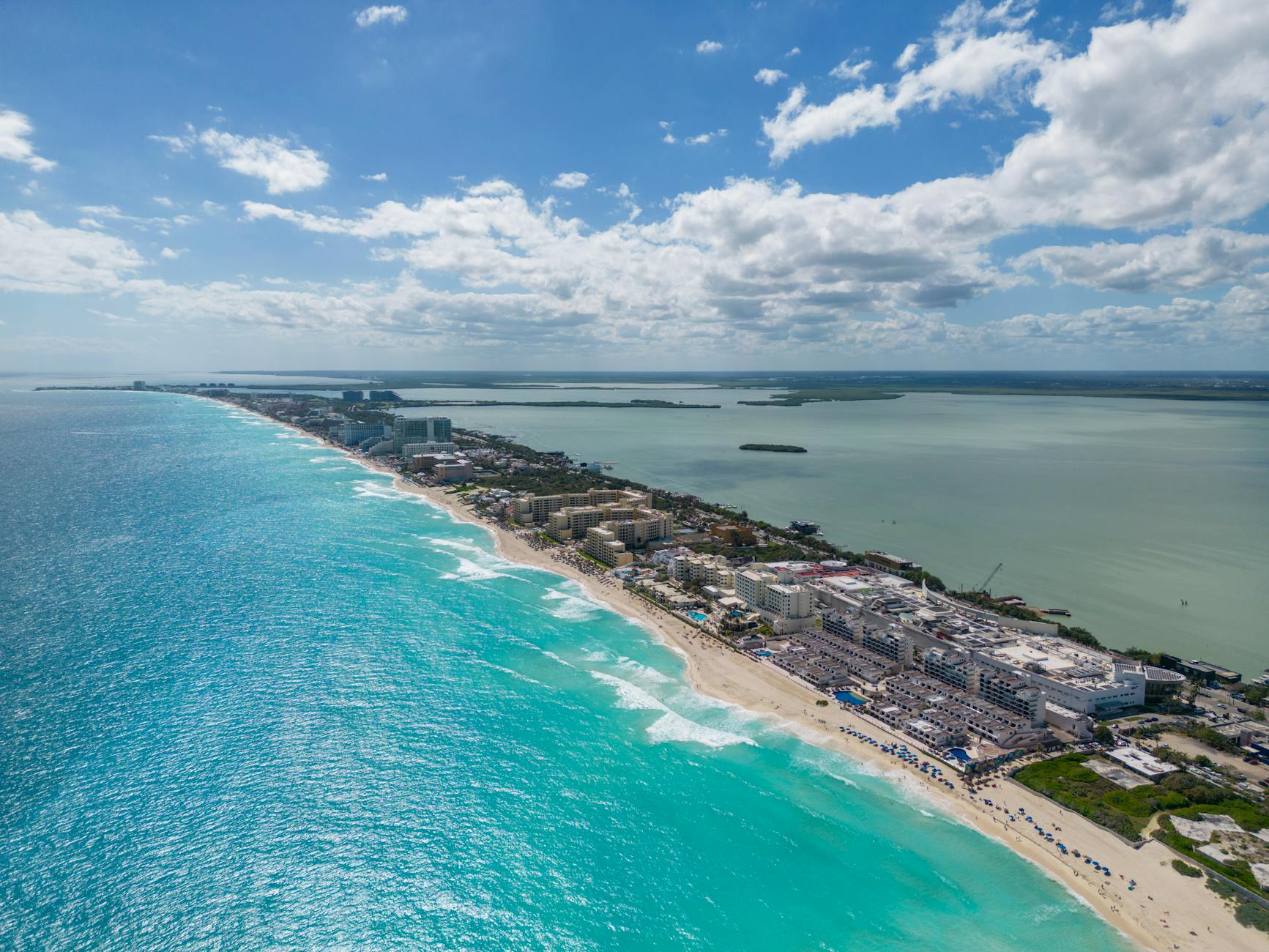Luftaufnahme einer langen Sandstrandpromenade mit Hotels und dem tuerkisblauen Meer.