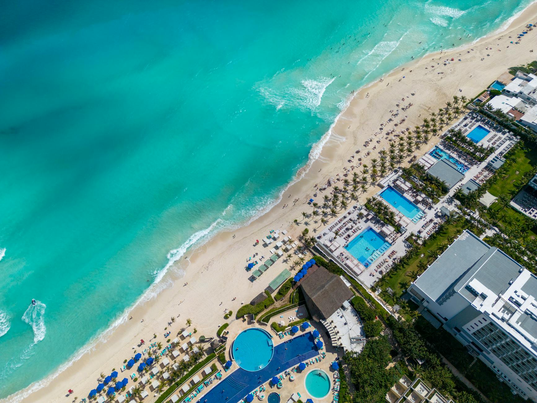 Luftaufnahme eines tropischen Sandstrandes mit tuerkisfarbenem Meer, Palmen und einer Hotelanlage mit mehreren Pools.