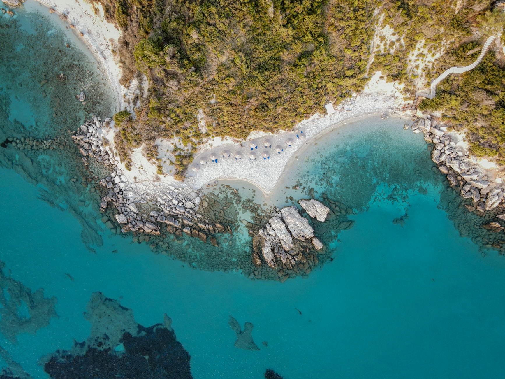 Chalkidiki, Griechenland - Luftaufnahme einer felsigen Bucht mit einem Sandstrand, an dem Sonnenschirme aufgestellt sind, und einem Holzweg, der sich ueber die Huegel schlauft.