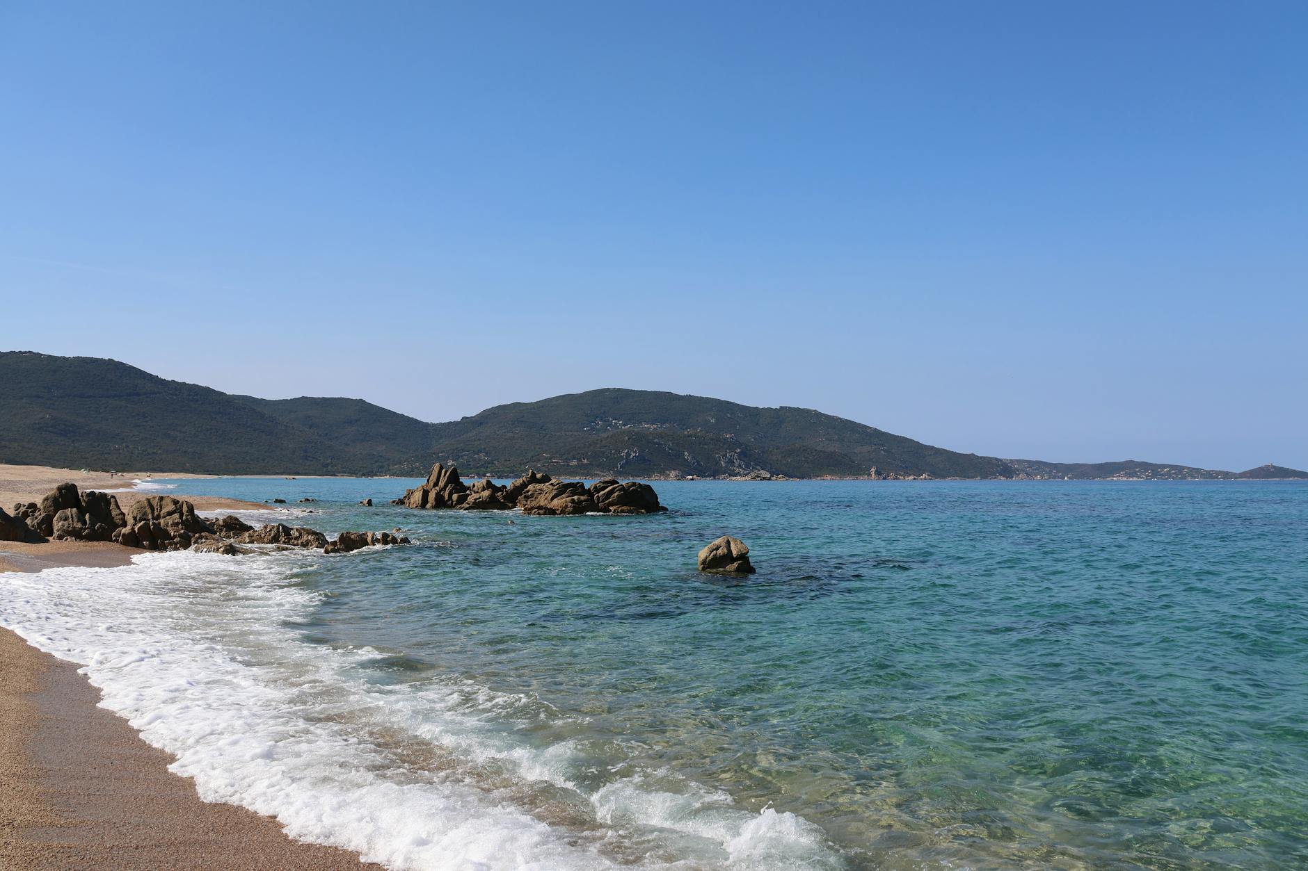 Chalkidiki, Griechenland - Ein Sandstrand mit Felsformationen im tuerkisen Meer und gruenen Huegeln im Hintergrund unter einem klaren blauen Himmel.
