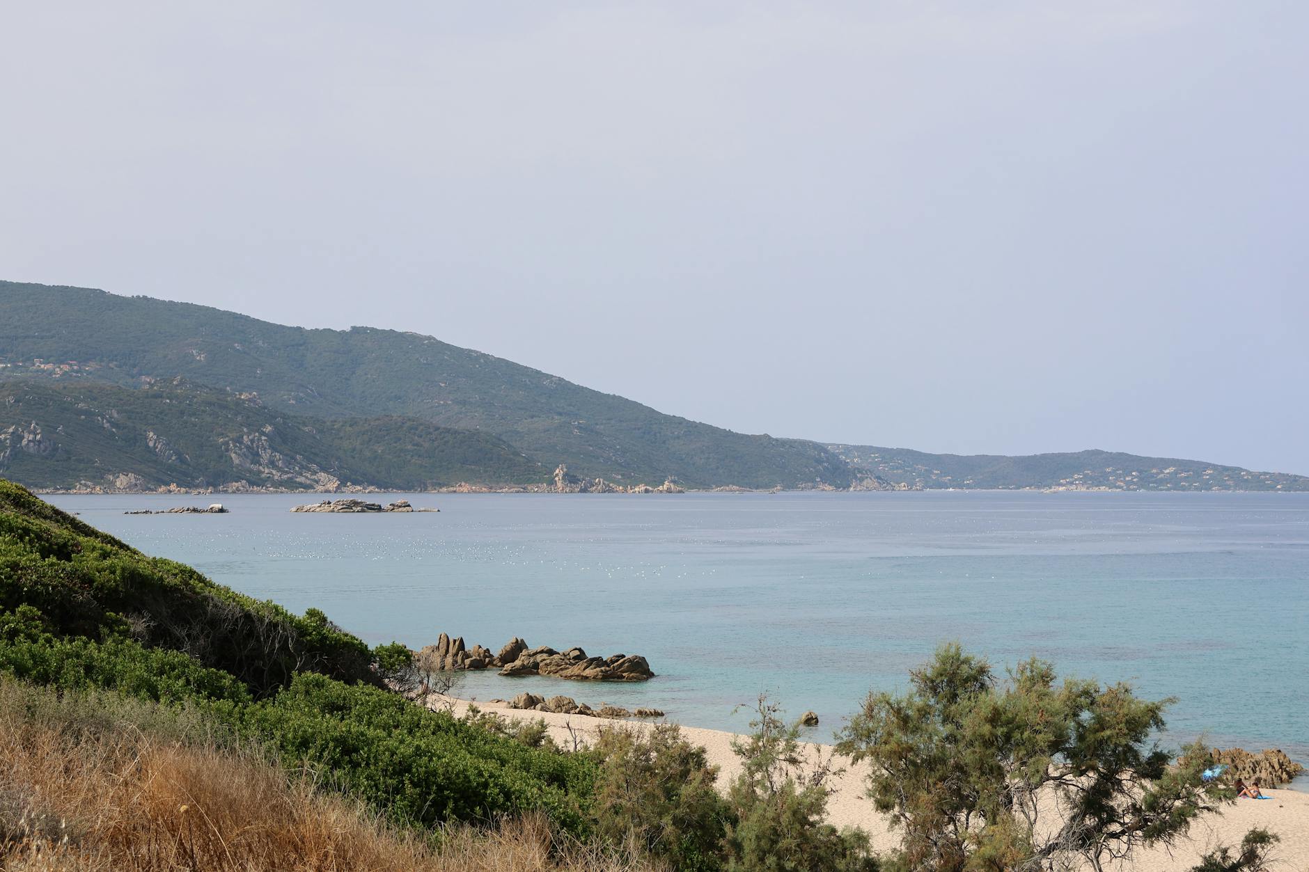 Chalkidiki, Griechenland - Ein Sandstrand mit felsigen Klippen und mediterraner Vegetation, der ins türkisfarbene Meer abfällt, mit bewaldeten Hügeln im Hintergrund.
