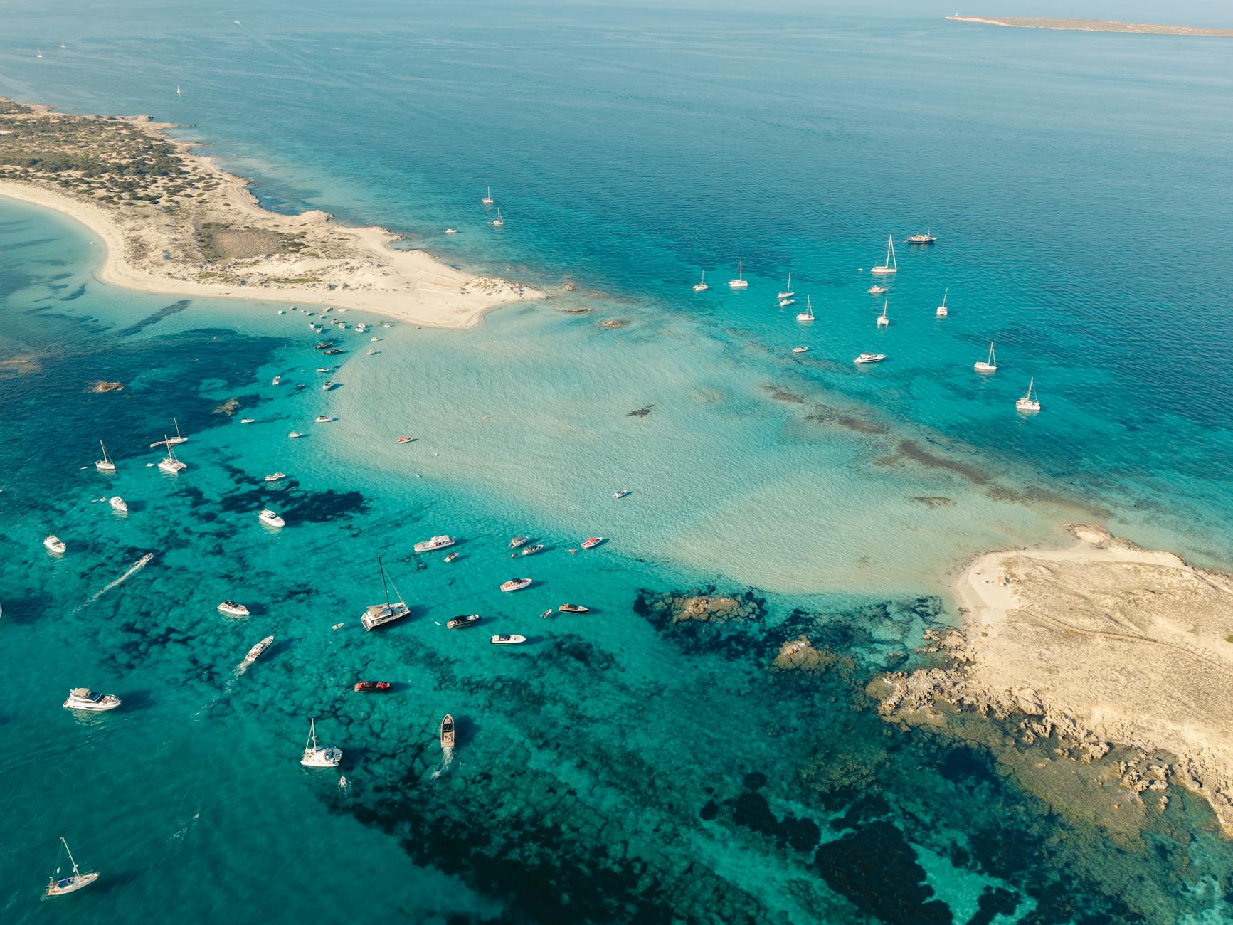 Luftaufnahme einer Küstenlinie mit vielen Booten und Segelbooten im türkisfarbenen Wasser.