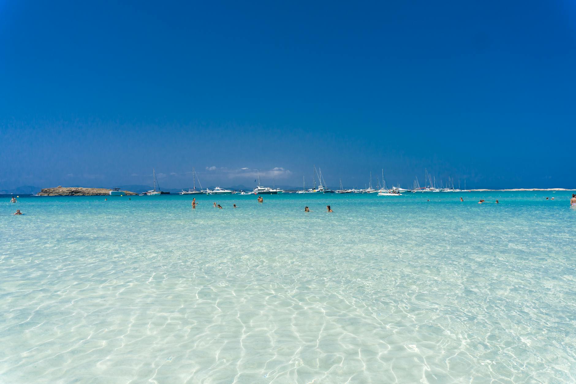 Menschen schwimmen in kristallklarem, türkisfarbenem Wasser vor einer Reihe von Segelbooten und Yachten, mit einer felsigen Insel am Horizont unter einem strahlend blauen Himmel.
