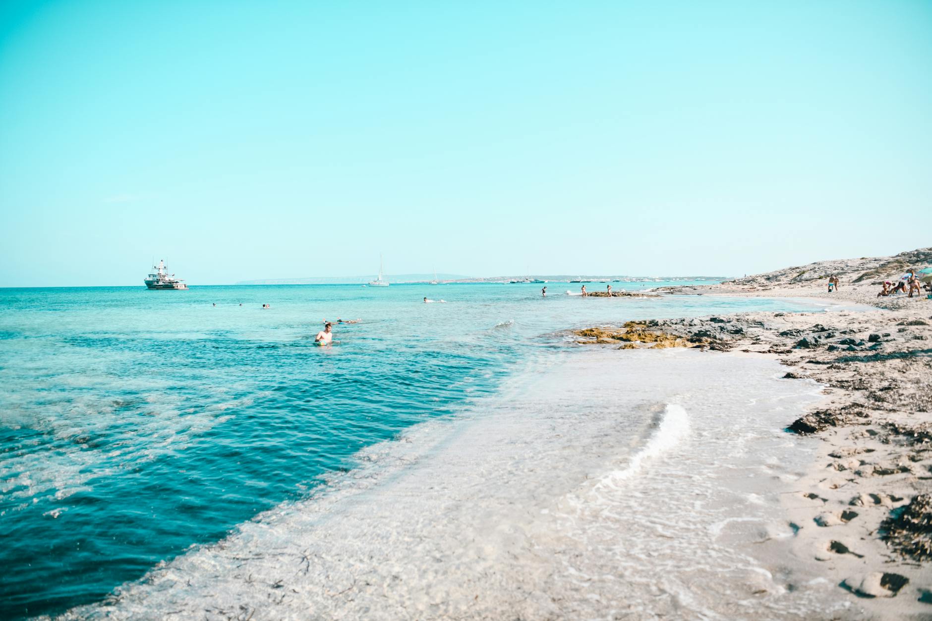 Ein sonniger Tag an einem felsigen Sandstrand mit klarem, tuerkisem Wasser und einigen Menschen, die schwimmen und sich am Ufer aufhalten.
