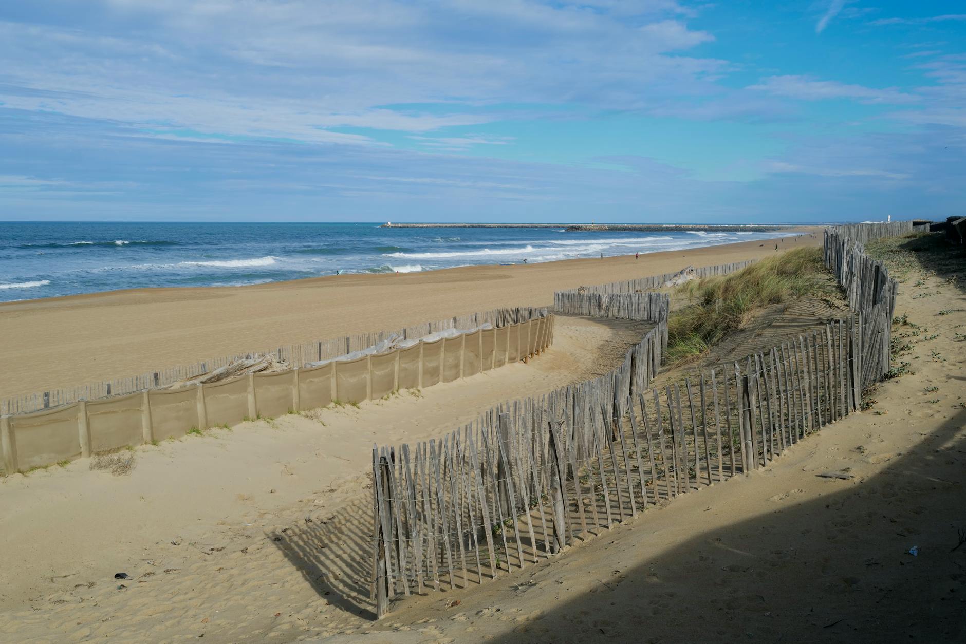 Ein weiter Sandstrand mit Wellen am Meer, gesäumt von Holzzäunen und Dünen.