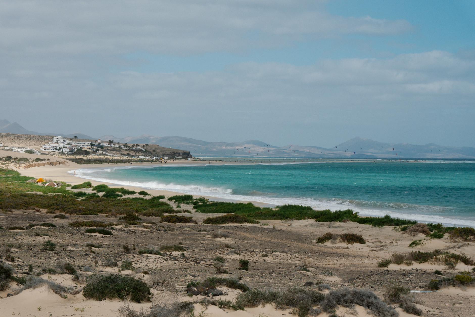 Ein weiter Sandstrand mit tuerkisblauem Wasser und vielen Kitesurfern, im Hintergrund eine duenenartige Landschaft mit kleinen Haeusern.