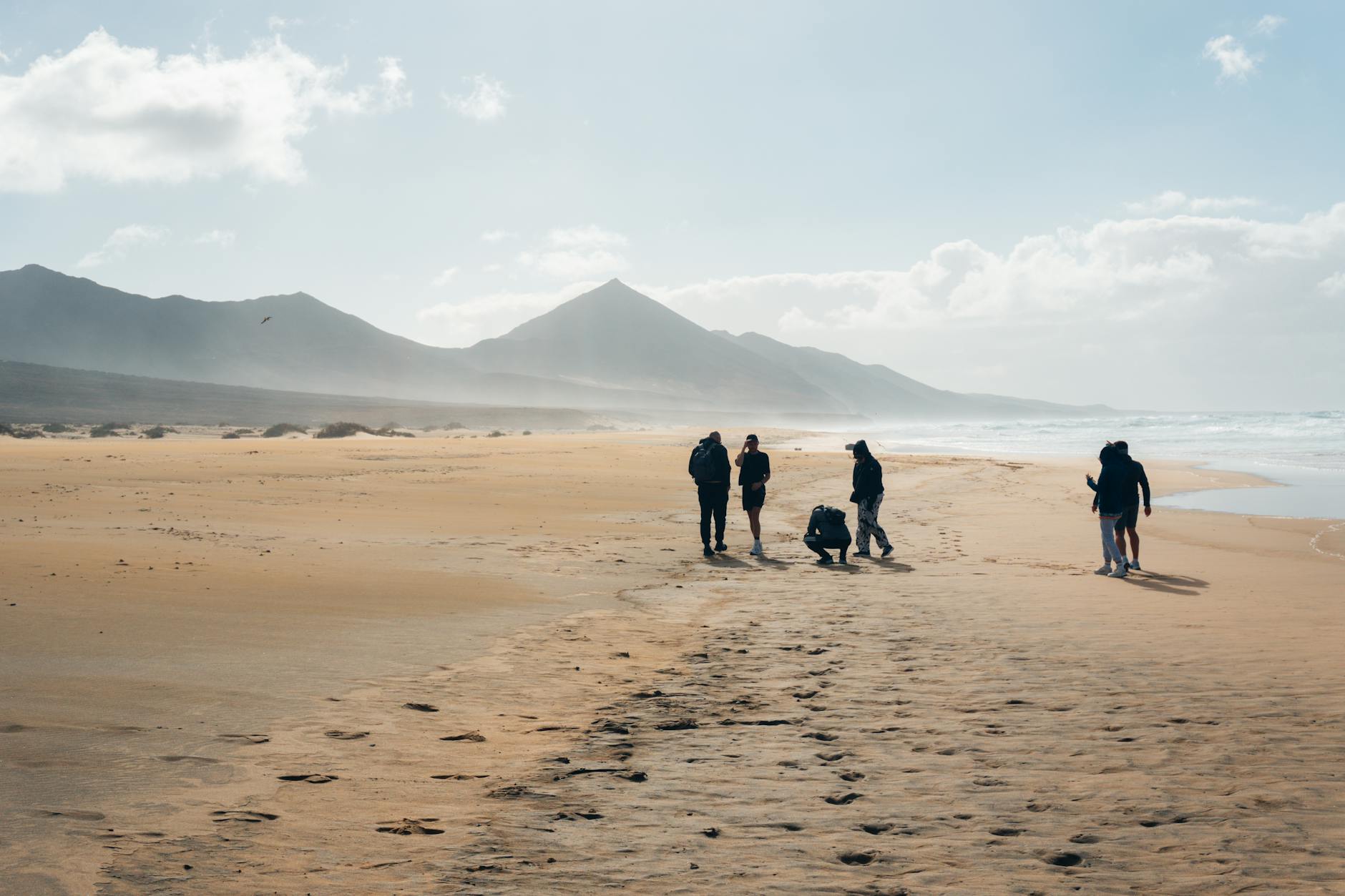 Eine Gruppe von Menschen geht auf einem weiten Sandstrand entlang, im Hintergrund sind Berge und das Meer zu sehen.