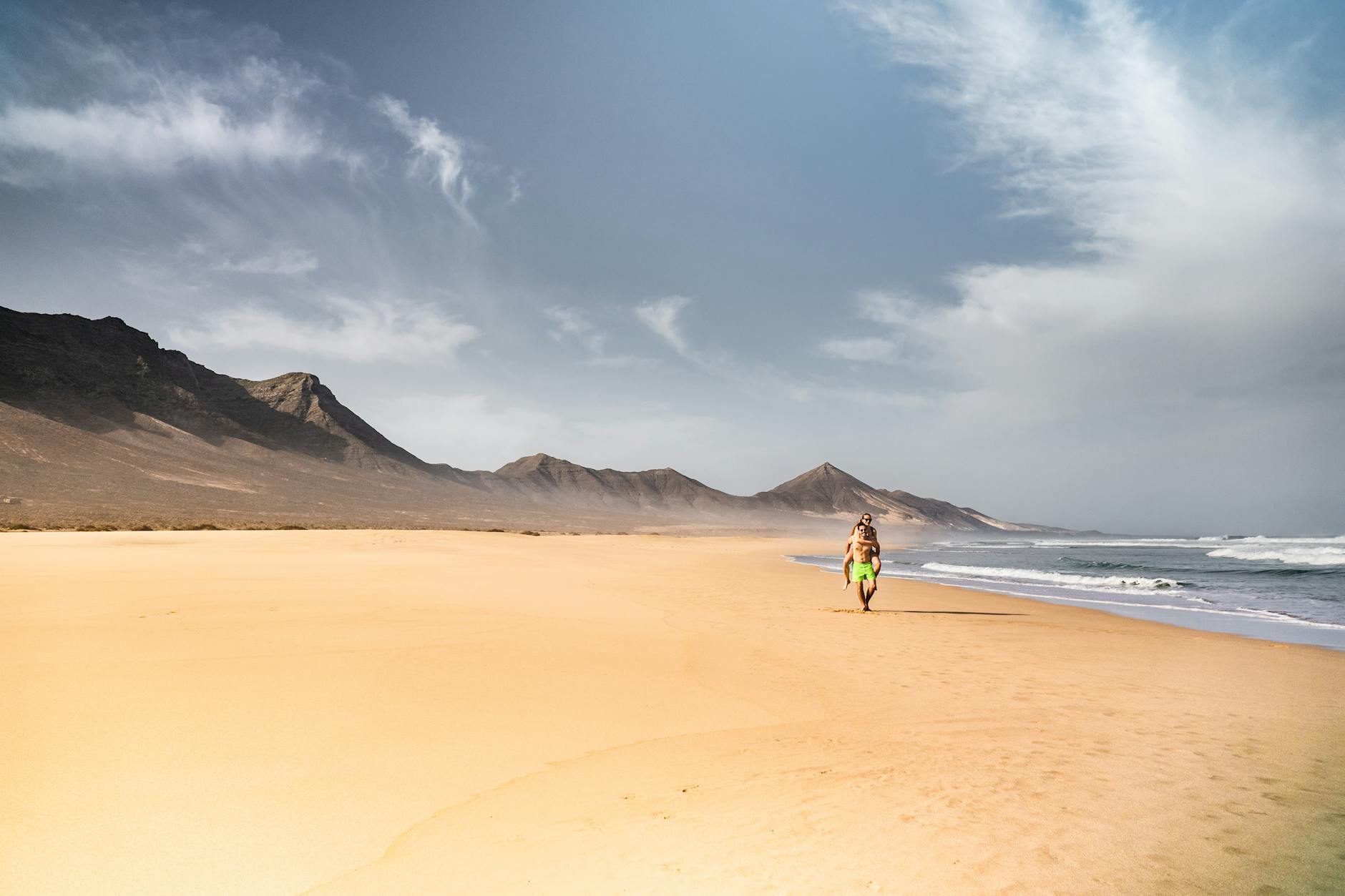 Ein Mann trägt eine Frau auf dem Rücken, während sie an einem breiten Sandstrand entlanggehen, mit Bergen im Hintergrund und Wellen, die an die Küste rollen.