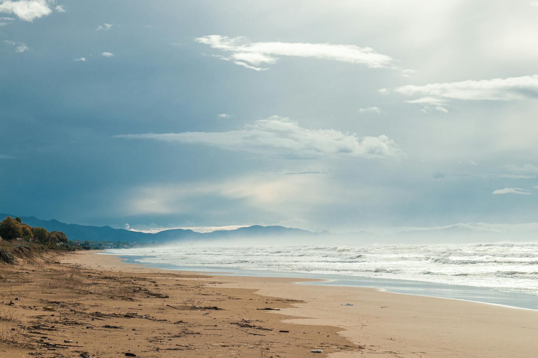 Ein weiter Sandstrand mit Blick auf das Meer und die Berge in der Ferne unter einem bewölkten Himmel.