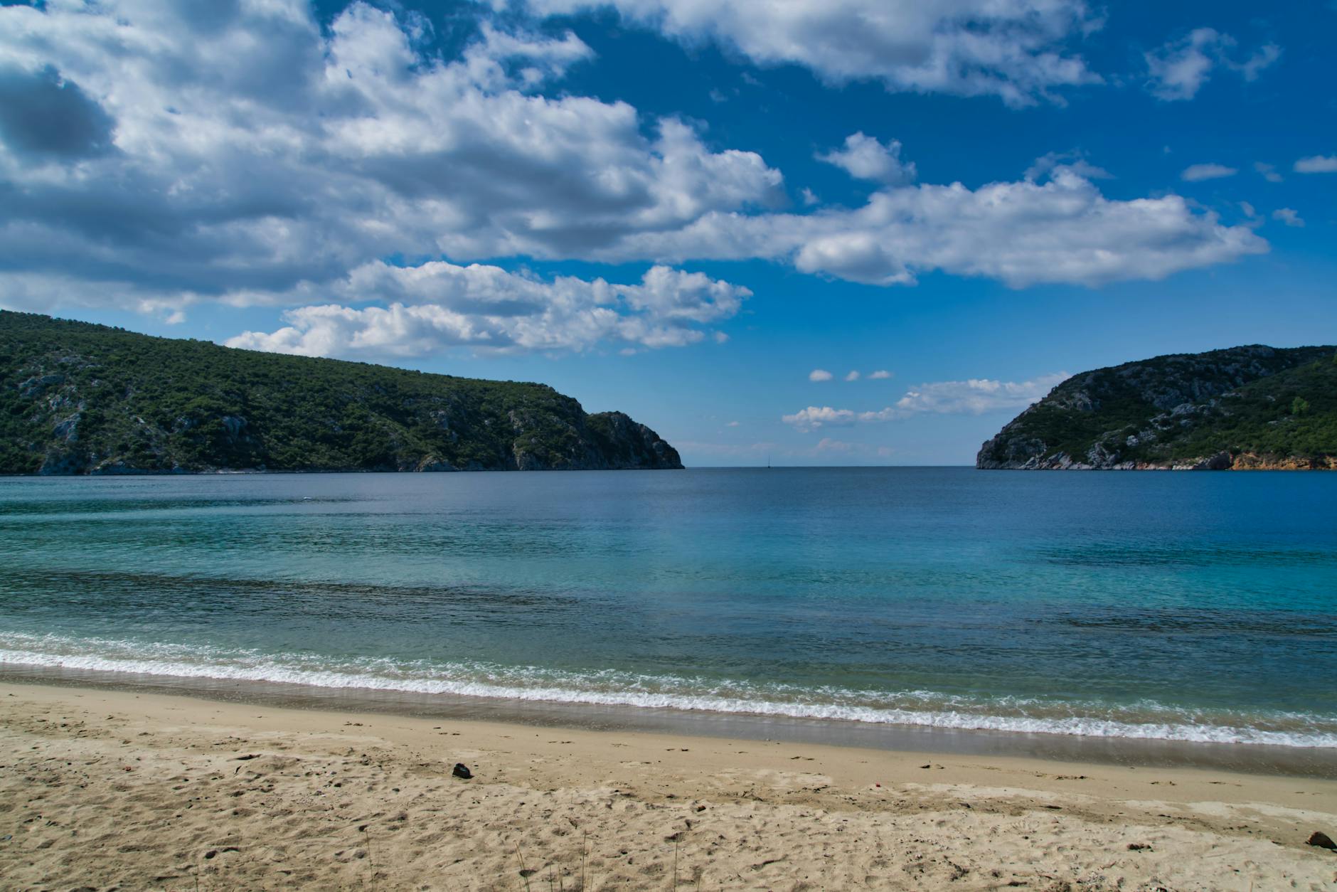 Ein Sandstrand mit tuerkisblauem Wasser und bewachsenen Huegeln im Hintergrund unter einem blauen Himmel mit Wolken.