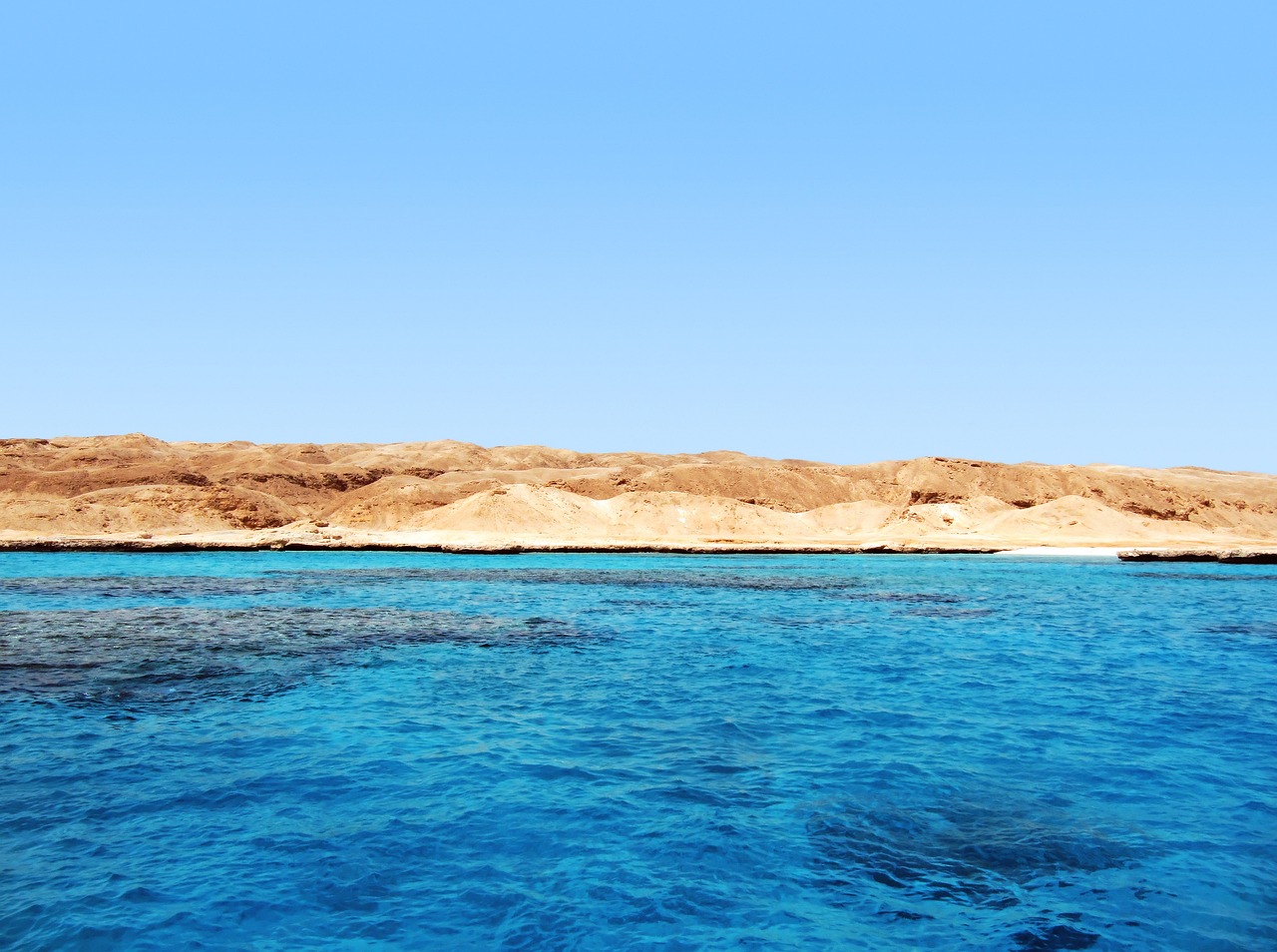 Ein Blick auf das tuerkisblaue Meer mit einer felsigen Wuestenlandschaft im Hintergrund unter einem klaren blauen Himmel.