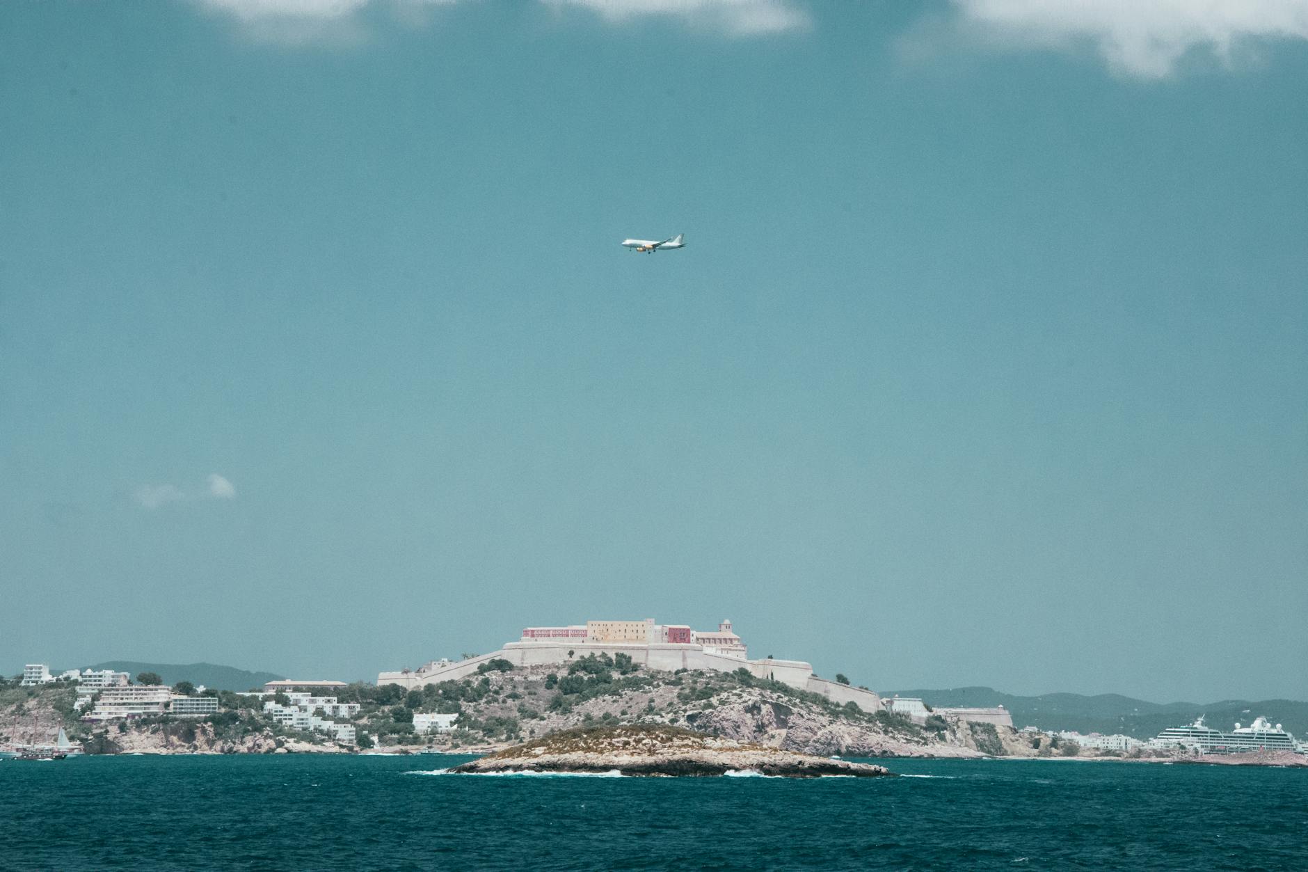 Ein Flugzeug fliegt am Himmel über einer Küstenstadt mit einer Festung auf einem Hügel und einem Kreuzfahrtschiff im Meer.