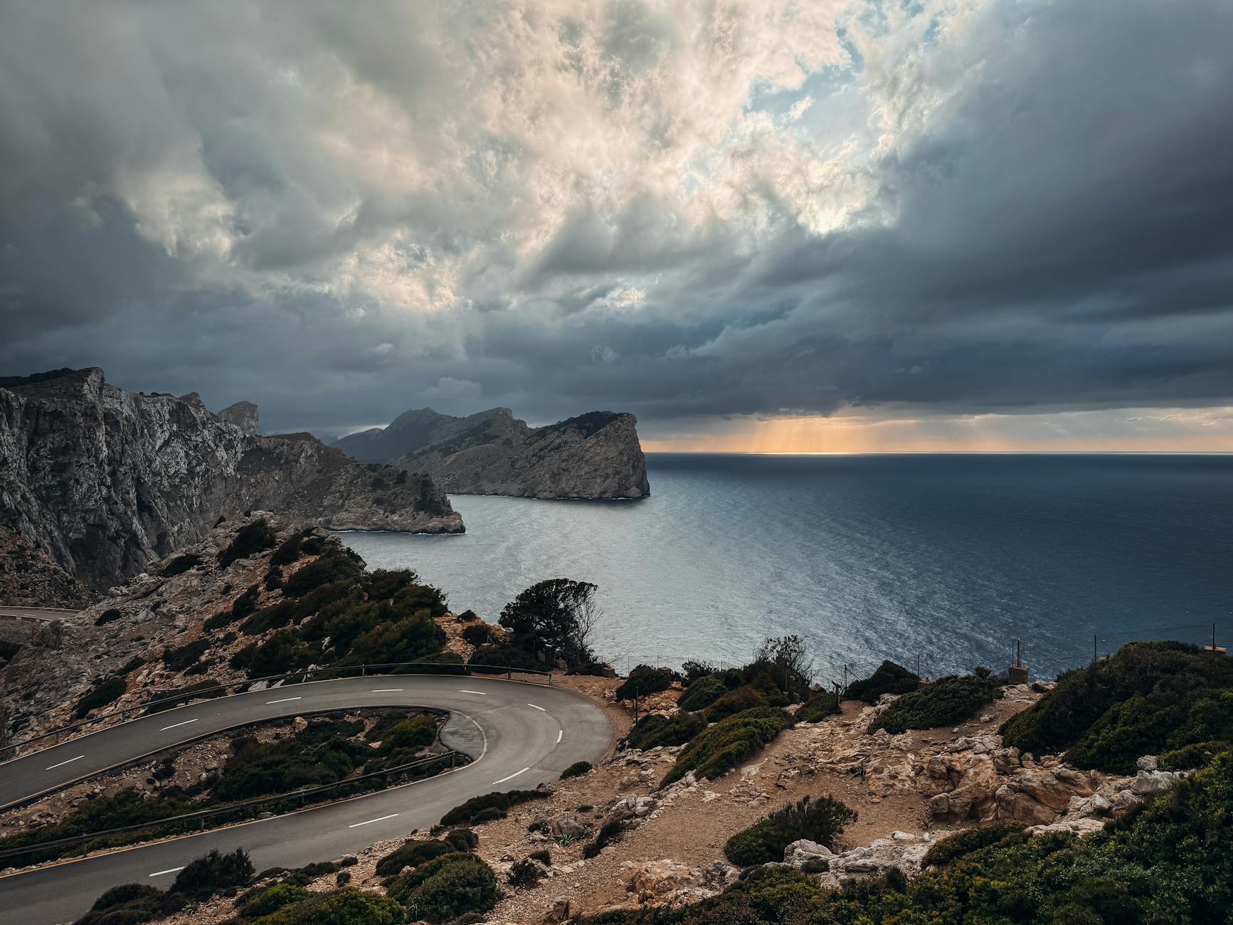 Eine kurvenreiche Küstenstraße führt über felsiges Gelände mit Blick auf das Meer und dramatische Wolkenformationen am Himmel.