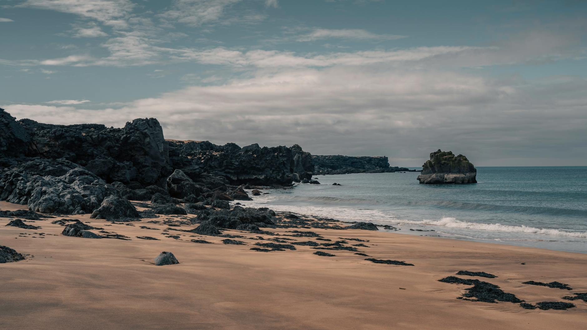 Ein sandiger Strand mit dunklen Felsformationen und dem Meer im Hintergrund unter einem bewölkten Himmel.