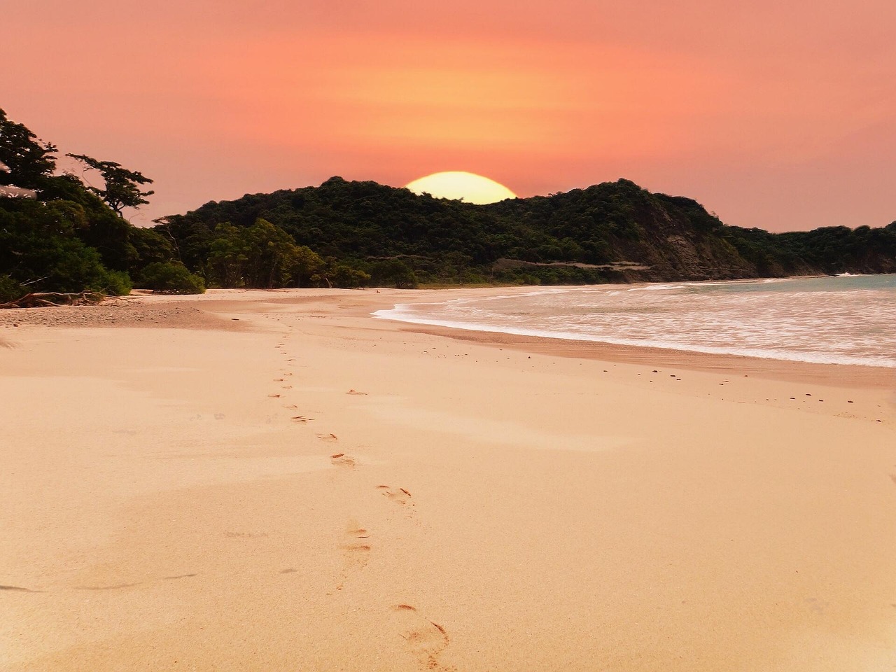 Ein Sandstrand mit Fussspuren im Vordergrund und einem Sonnenuntergang ueber dem Meer und einer bewaldeten Kueste im Hintergrund.