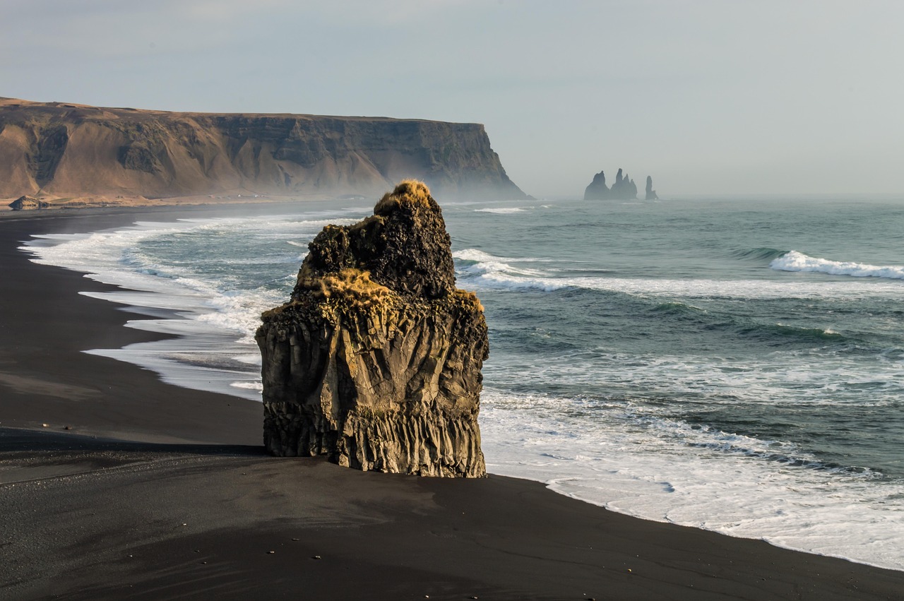 Ein einzelner, moosbewachsener Felsen steht auf einem schwarzen Sandstrand am Meer, mit Wellen, die an die Kueste rollen.