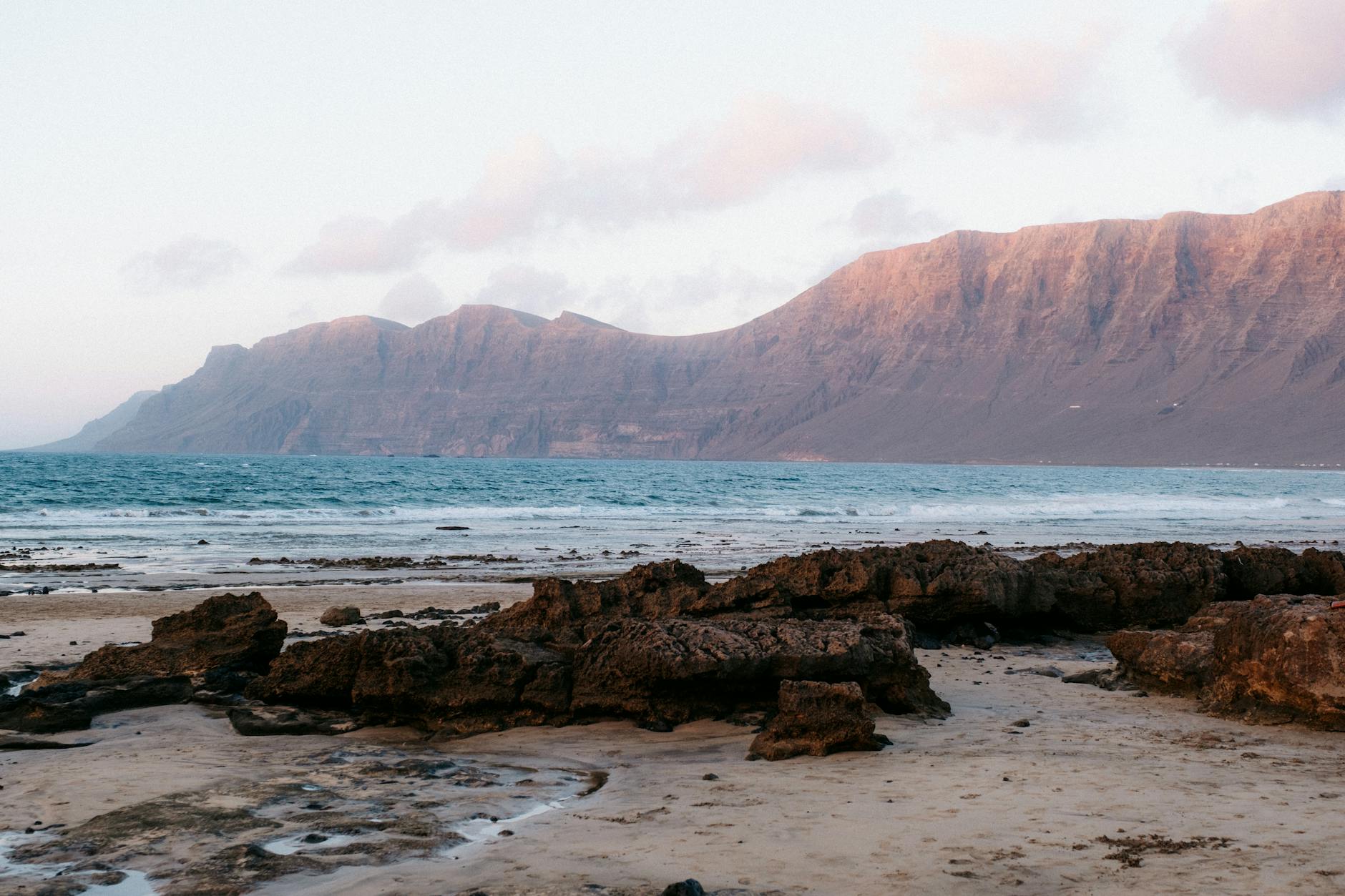 Kanaren, Spanien - Ein felsiger Strand mit Wellen, die auf das Ufer treffen, und einer Bergkette im Hintergrund.
