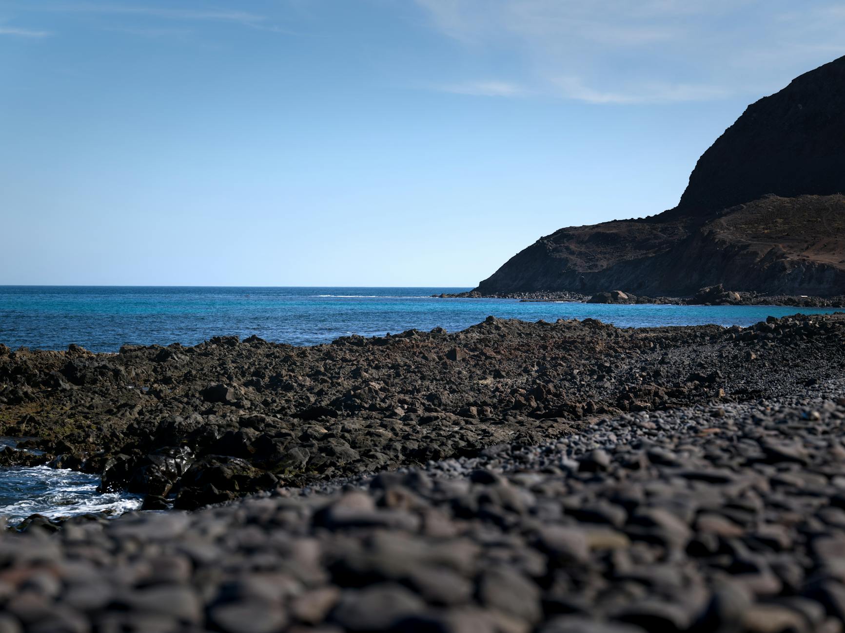 Kanaren, Spanien - Ein felsiger Strand mit dunklen, vulkanischen Felsen im Vordergrund und dem tuerkisblauen Meer, das sich bis zu einer dunklen Bergkueste erstreckt.