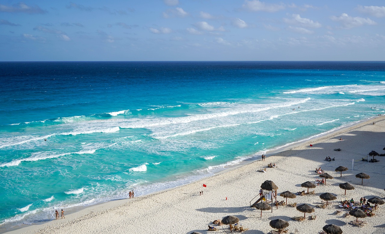 Ein weiter Sandstrand mit tuerkisblauem Meer und Wellen, im Vordergrund Sonnenschirme und einige Menschen.