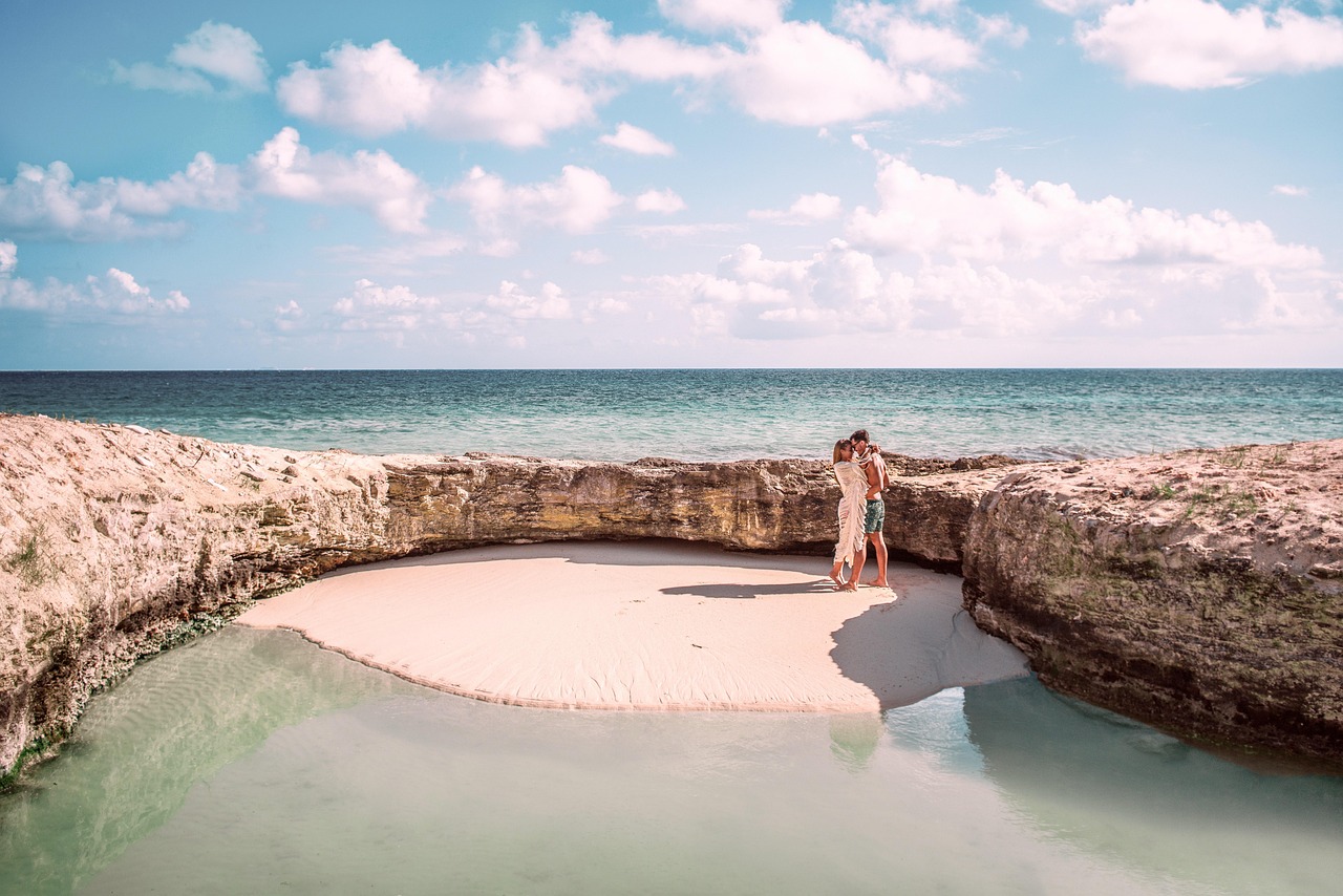 Ein Paar steht auf einem sandigen Strandabschnitt, der von Felsformationen umgeben ist, mit dem Meer und einem bewölkten Himmel im Hintergrund.