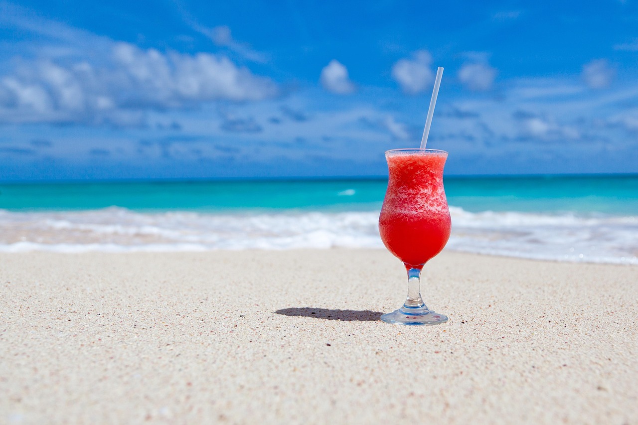 Ein roter Cocktail mit einem Strohhalm steht auf einem Sandstrand vor dem türkisfarbenen Meer und blauem Himmel mit Wolken.