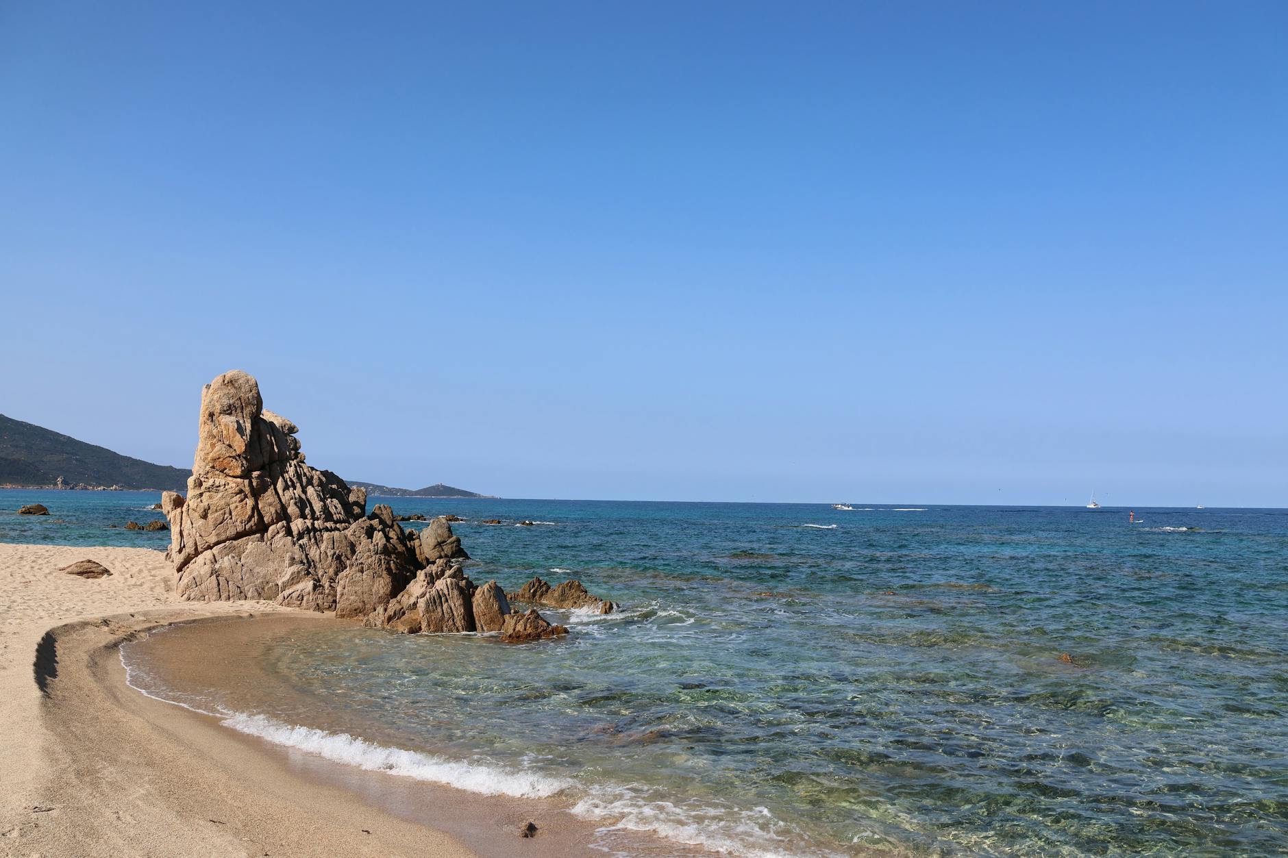 Ein sandiger Strand mit einer markanten Felsformation im Vordergrund, das klare, tuerkise Meer und Boote am Horizont unter einem blauen Himmel.