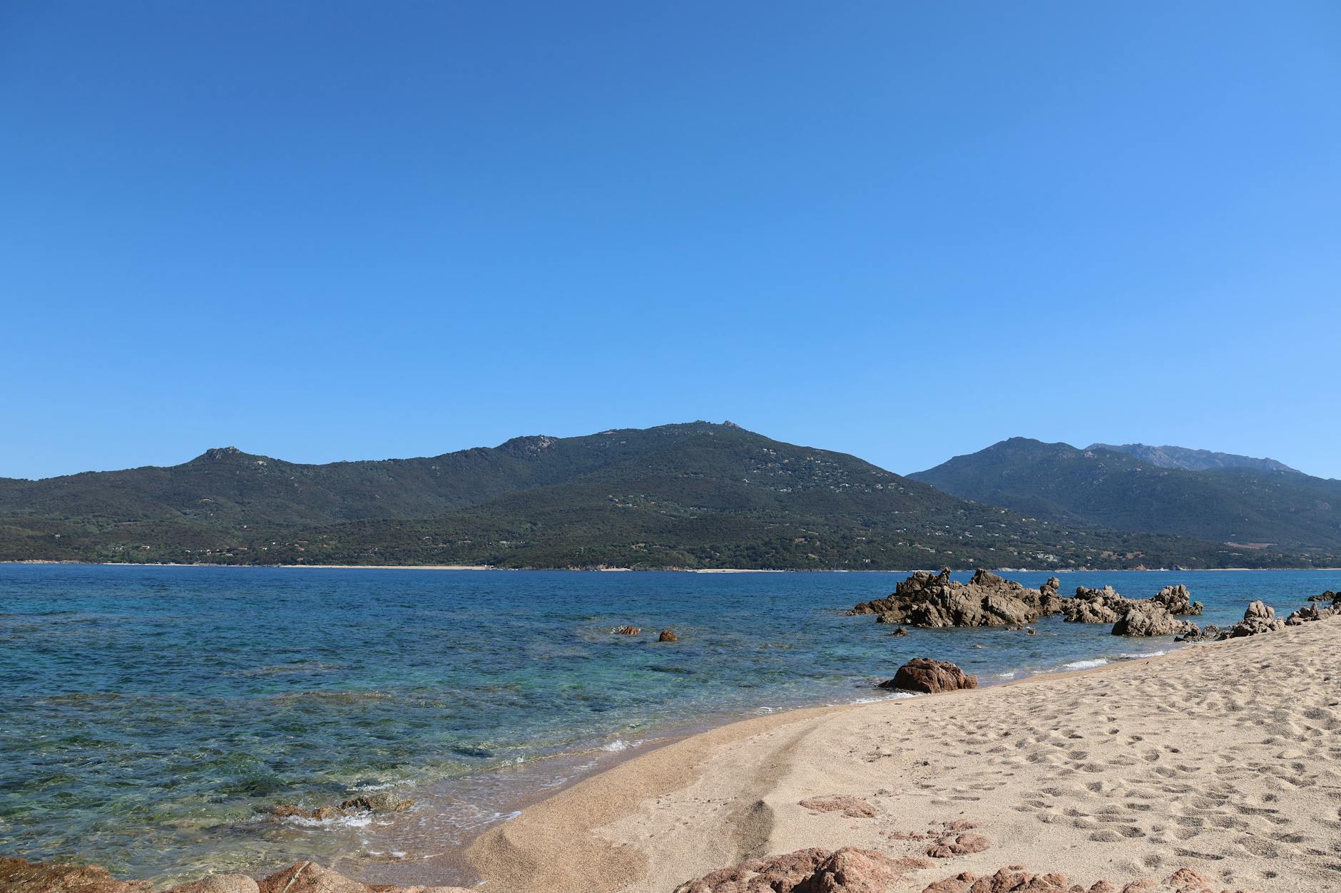 Ein sandiger Strand mit felsigen Klippen im türkisfarbenen Meer und bewaldeten Bergen im Hintergrund unter einem klaren blauen Himmel.