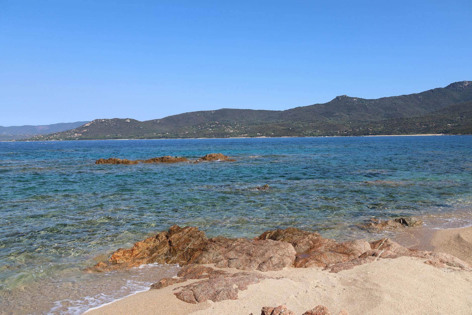 Ein Sandstrand mit felsigen Abschnitten und klarem, blauem Wasser, im Hintergrund eine bewaldete Bergkette.