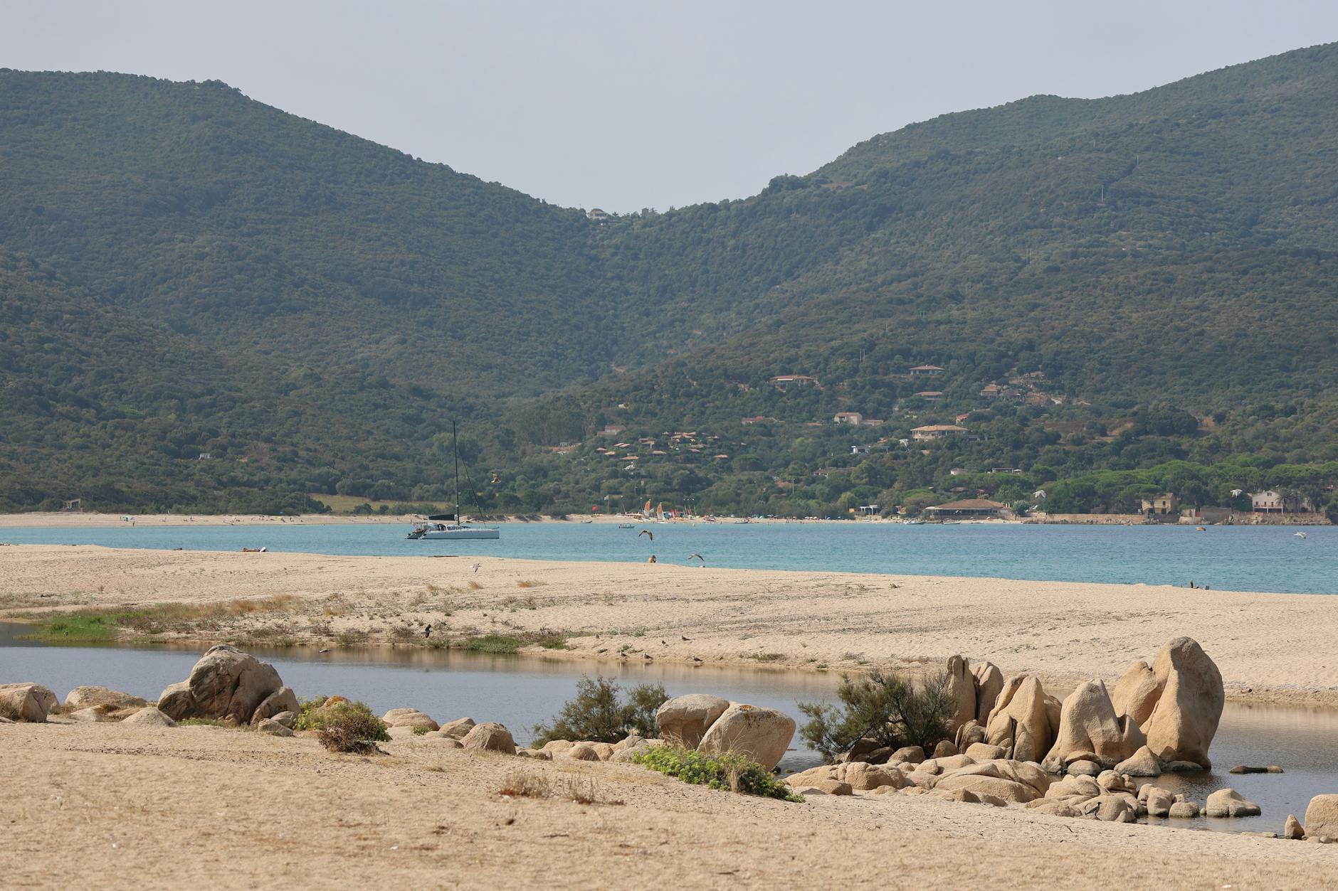 Ein sandiger Strand mit Felsformationen im Vordergrund, dahinter das blaue Meer mit einem Segelboot und bewaldeten Hängen.