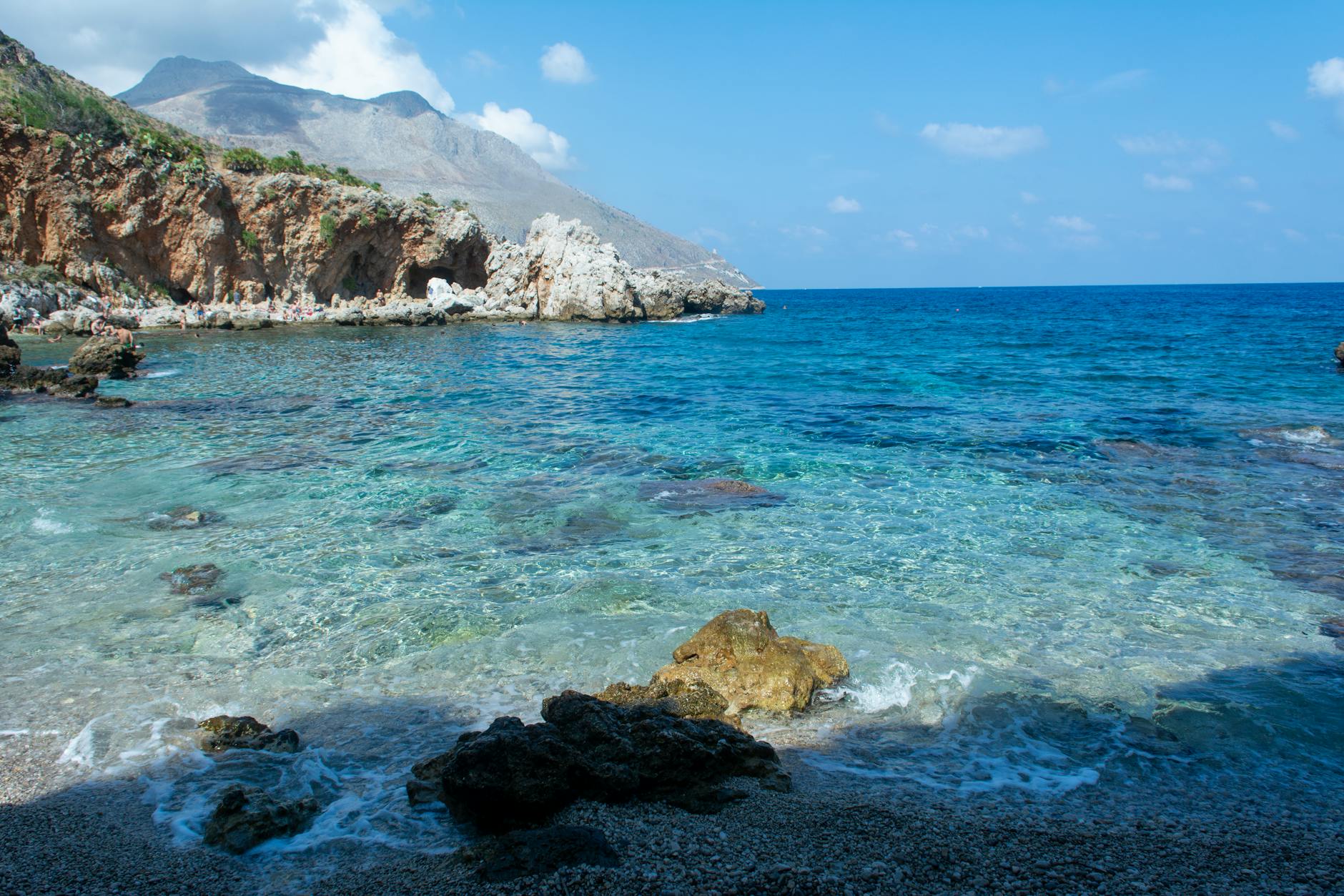 Ein Kiesstrand mit Felsen und klarem, türkisfarbenem Wasser, im Hintergrund eine felsige Küste mit Höhlen und Bergen.