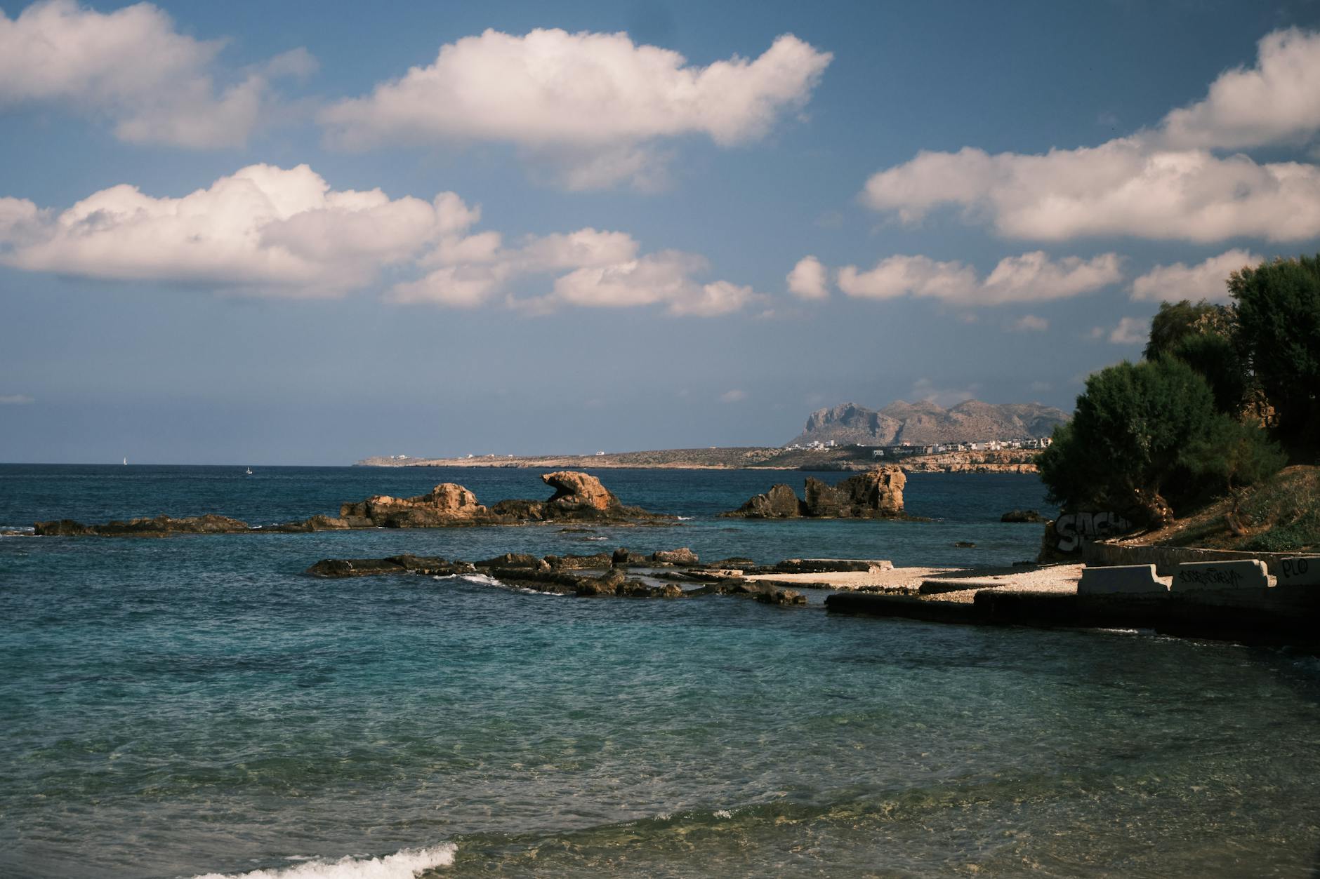 Eine felsige Küste mit klarem, türkisfarbenem Wasser und einigen Felsformationen im Meer, im Hintergrund eine Küstenstadt und Berge.