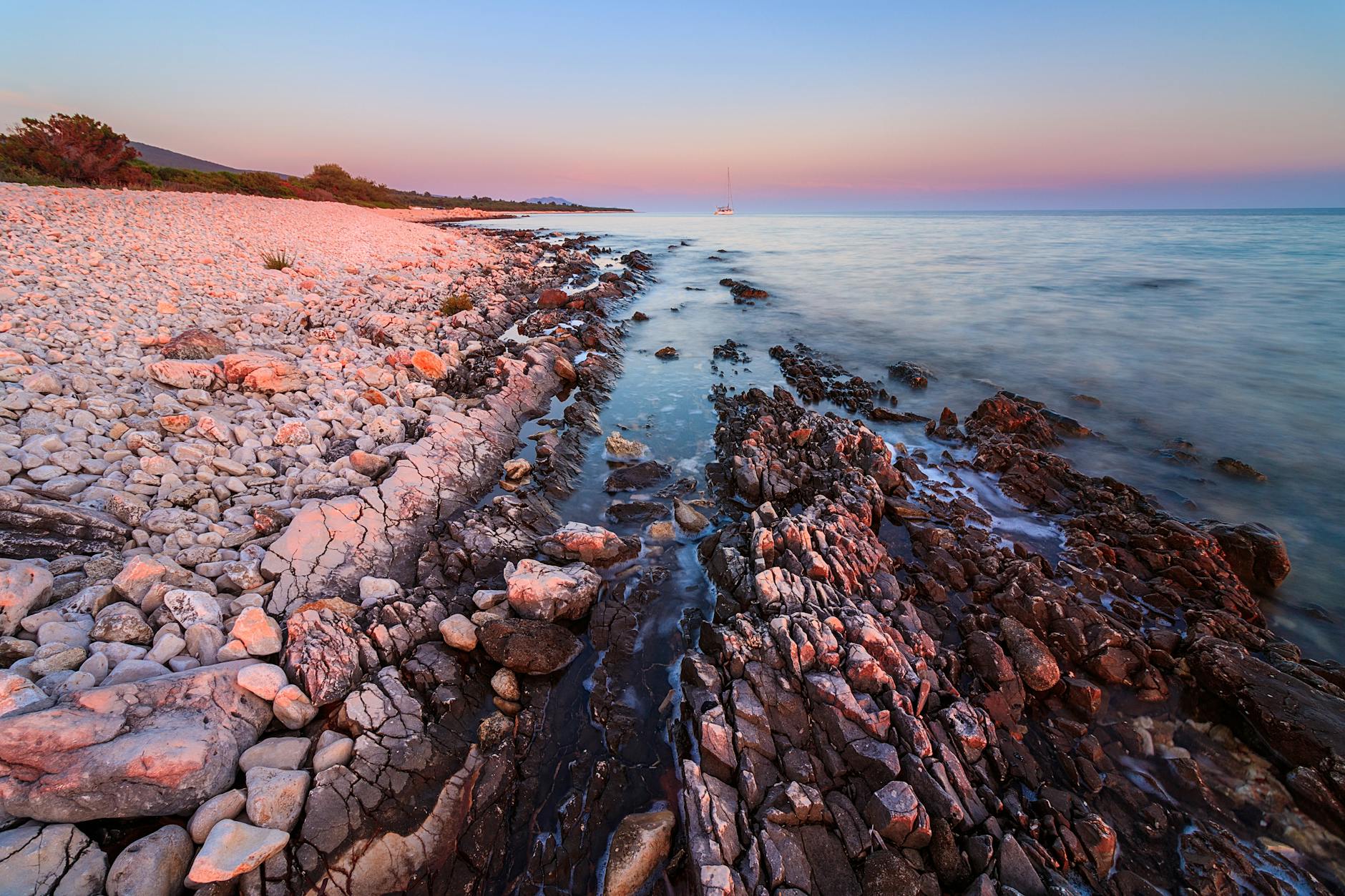 Ein Kiesstrand mit Felsen und dem Meer bei Sonnenuntergang, ein Segelboot ist in der Ferne zu sehen.