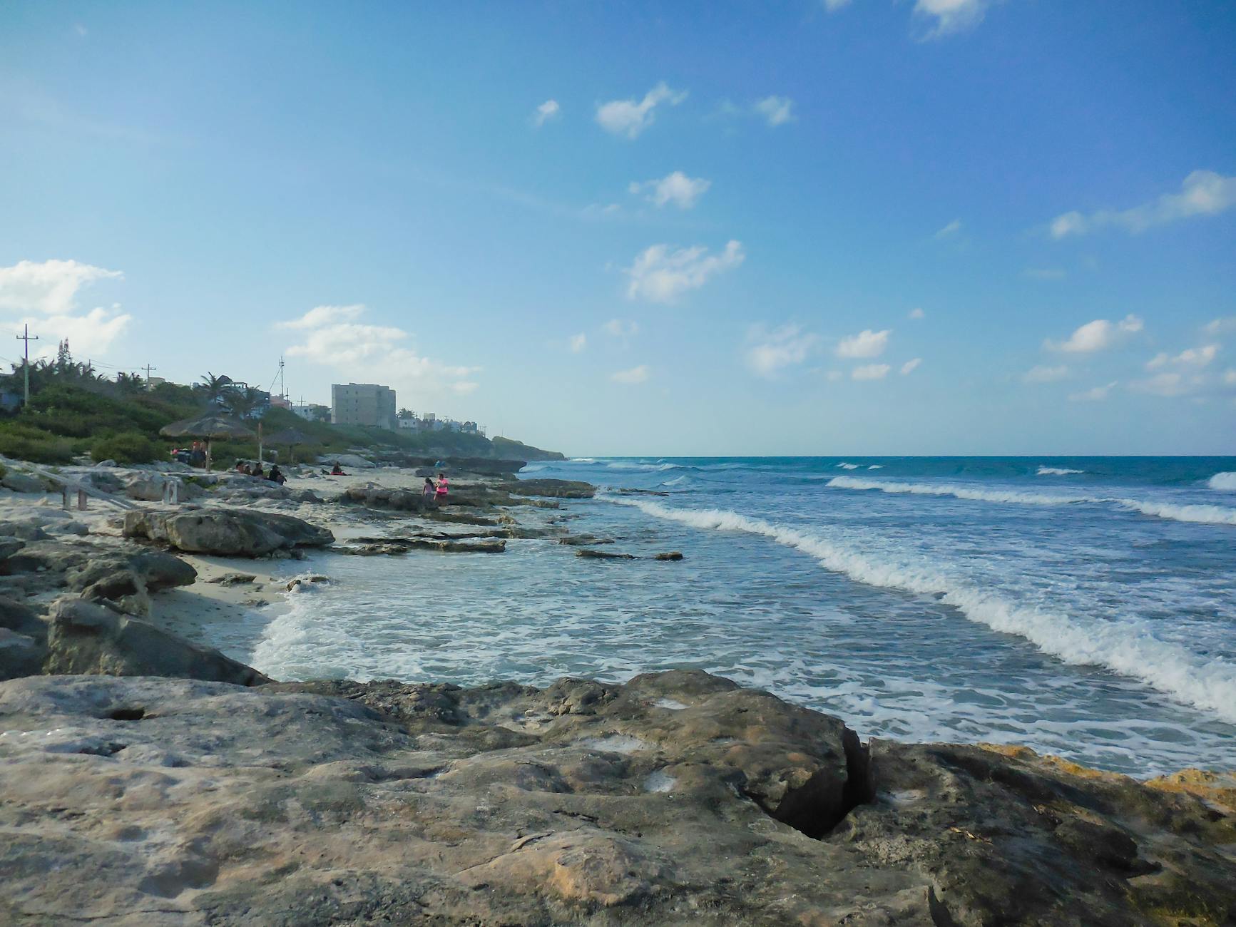 Ein felsiger Strand mit dem Meer und Wellen im Hintergrund, im Hintergrund sind Palmen und einige Gebäude zu sehen.