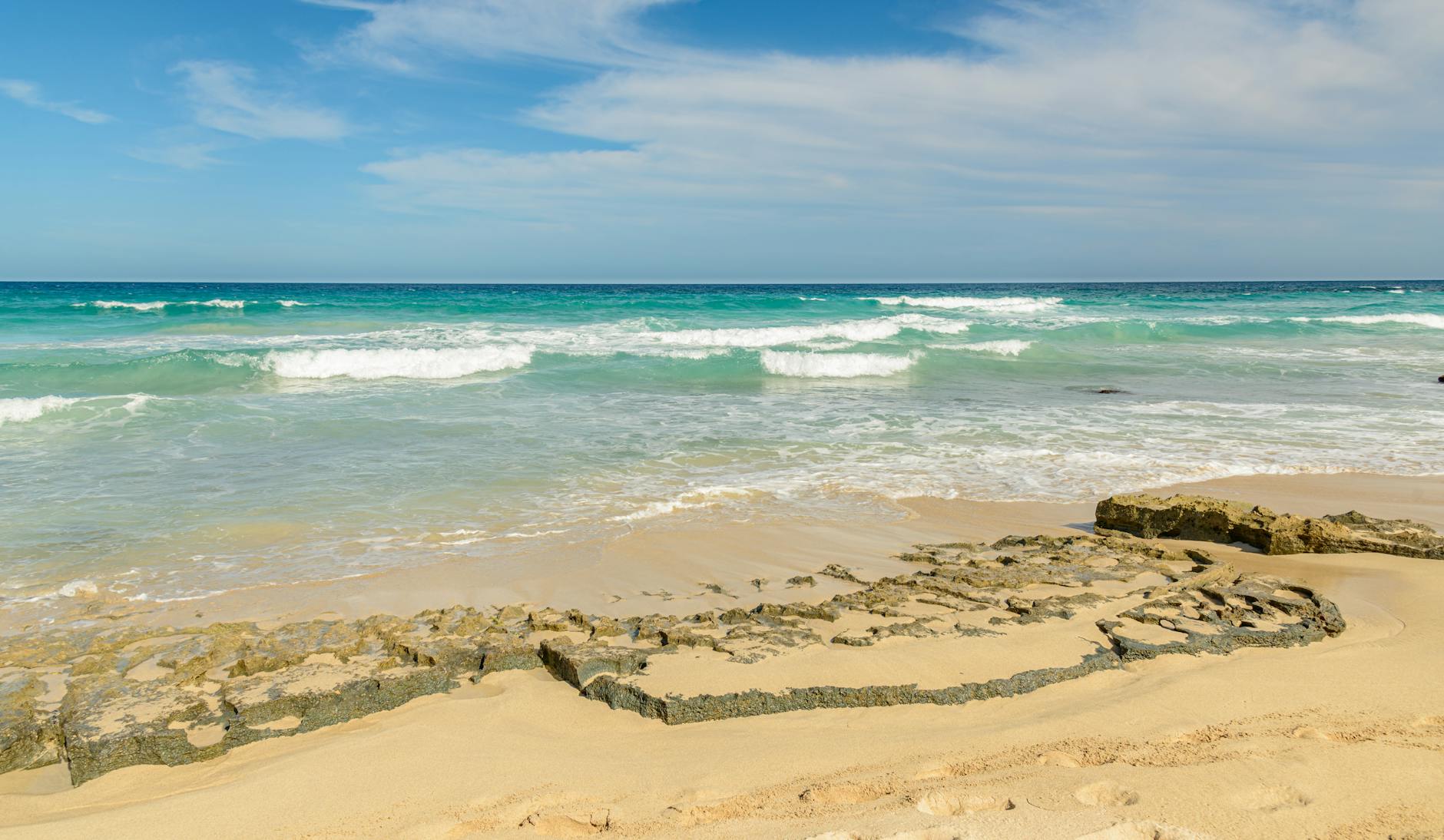 Ein Sandstrand mit felsigen Formationen im Vordergrund und dem tuerkisen Meer mit Wellen und blauem Himmel im Hintergrund.